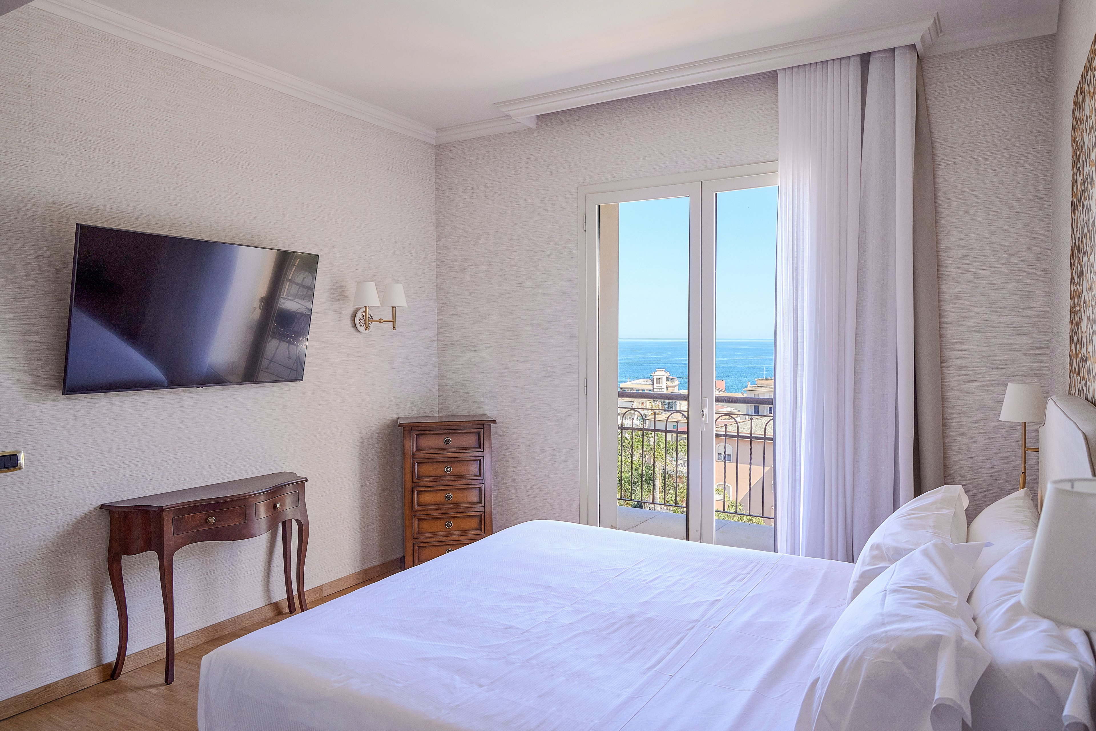 A sunlit hotel bedroom features a neatly made double bed with white linens facing a wall-mounted flat-screen TV. French doors open onto a balcony overlooking buildings and a distant blue sea under a clear sky. A small wooden desk and tall chest of drawers add classic touches to the otherwise modern, minimal decor. Soft neutral walls and light curtains create a calm, airy atmosphere ideal for relaxation.