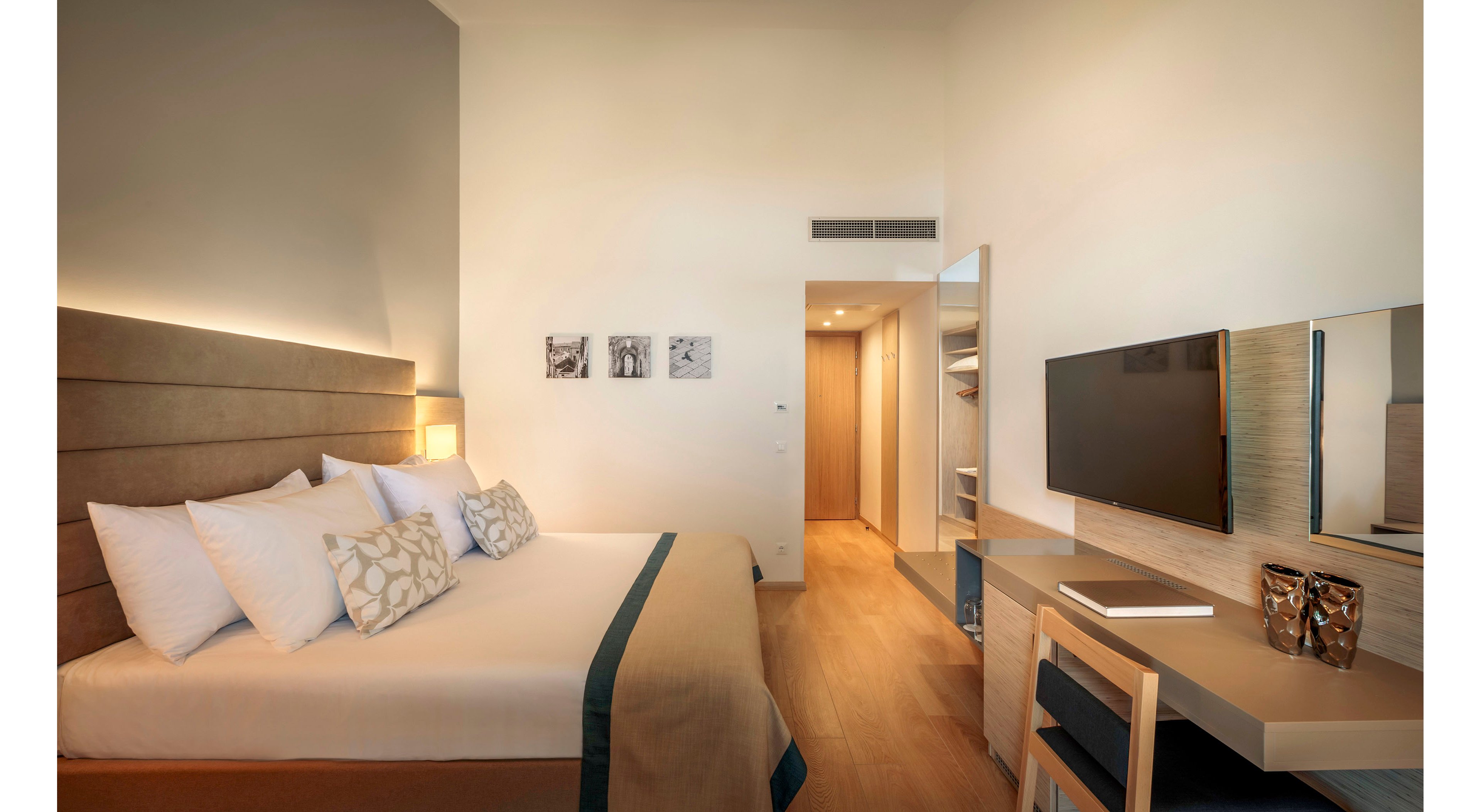 A contemporary hotel room features a large bed with crisp white linens and patterned accent pillows. The space includes a sleek wooden desk with a flat-screen television mounted on the wall and a mirror above the work area. Warm recessed lighting and neutral tones create a calm, inviting atmosphere. A hallway with built-in storage leads toward the entrance, enhancing the room’s clean, minimalist aesthetic.