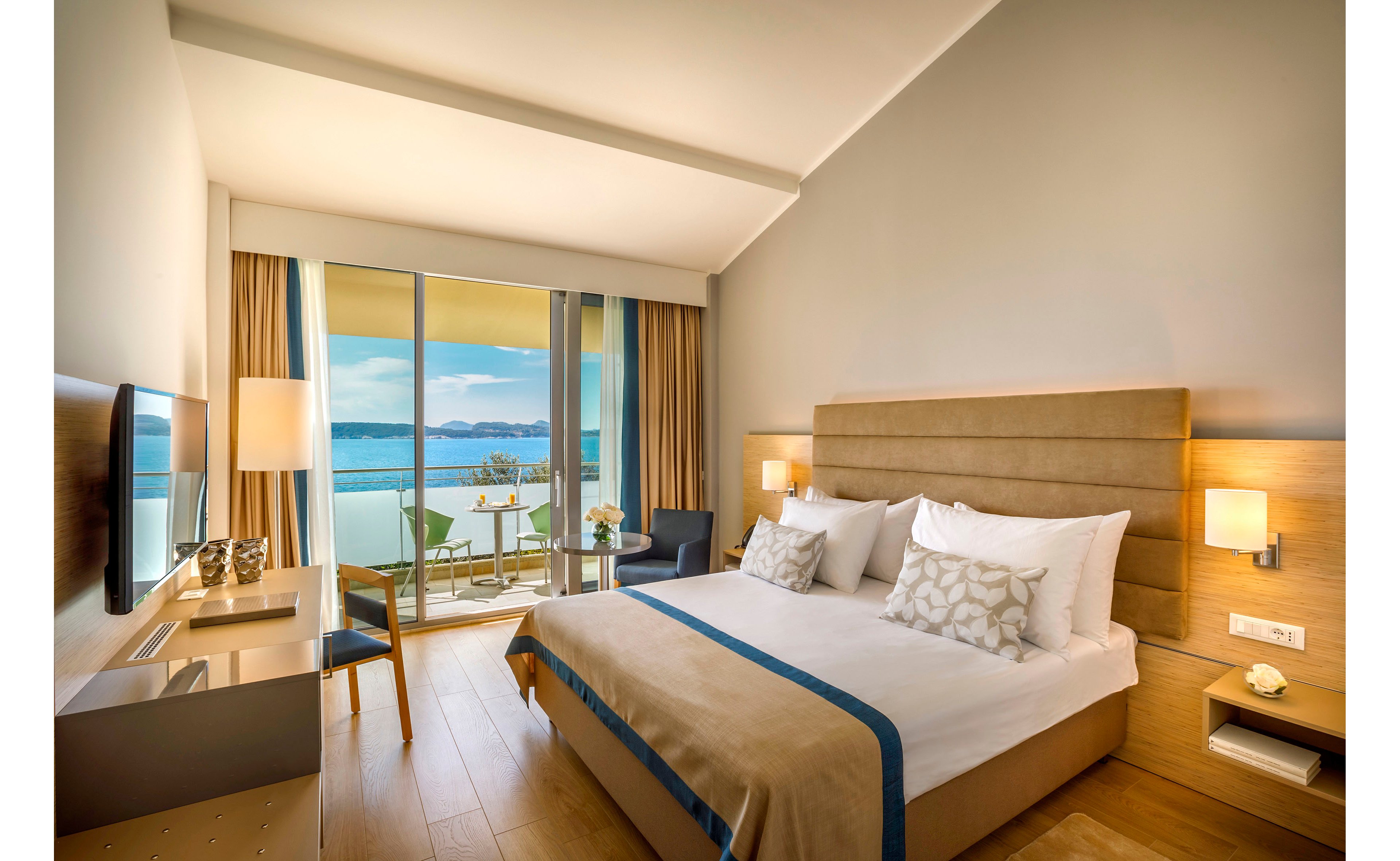 A contemporary hotel bedroom features a large double bed with neutral bedding and decorative pillows. Floor-to-ceiling glass doors open onto a balcony overlooking a calm sea and distant coastline. The room includes a desk with a flat-screen TV, a small seating area, and warm wooden flooring. Soft lighting from wall lamps and a table lamp creates a cozy, inviting atmosphere.