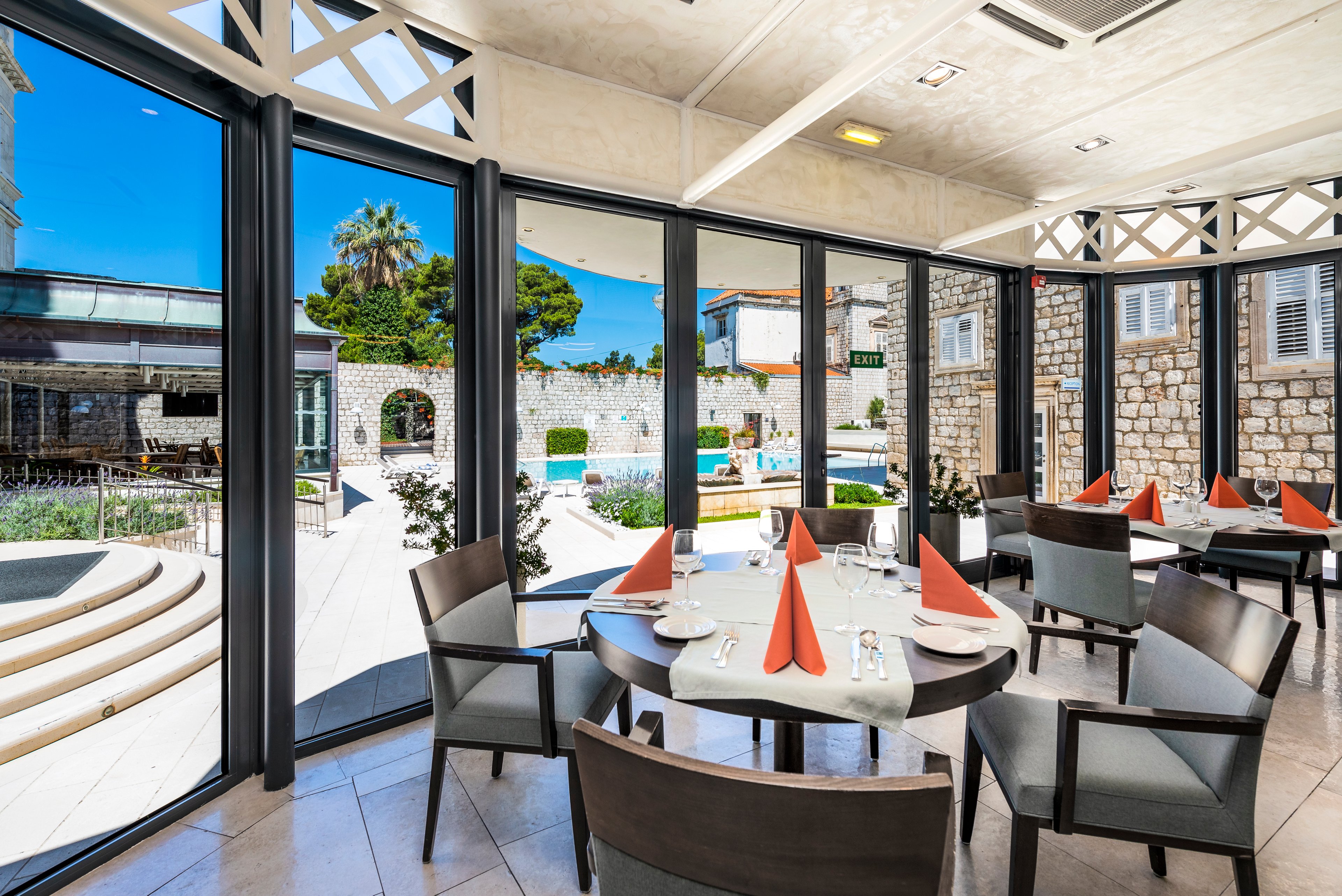 A sunlit restaurant dining area features round tables set with white tablecloths and orange folded napkins. Large floor-to-ceiling windows provide a clear view of an outdoor swimming pool and stone courtyard. Modern chairs surround the tables, and glassware is neatly arranged, suggesting readiness for guests. The scene conveys a relaxed, upscale atmosphere in a resort or hotel setting.