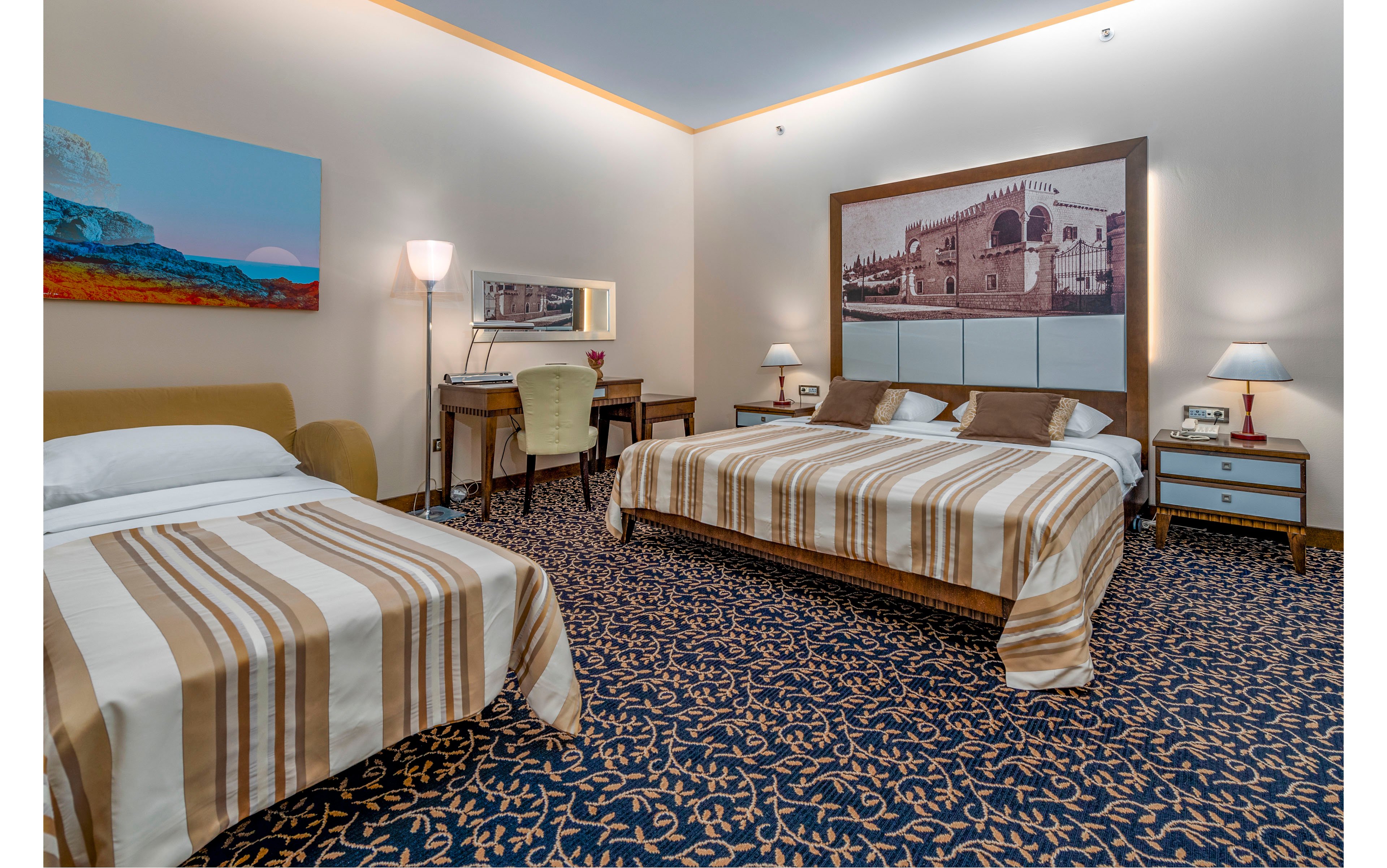 A spacious hotel room features a large double bed and a smaller single bed, both dressed in striped beige and brown bedding. The room includes bedside tables with lamps, a writing desk with a mirror, and a cushioned chair. Wall art above the headboard and on the side wall adds visual interest, while patterned carpet covers the floor. Soft, warm lighting creates a comfortable and inviting atmosphere suitable for guests.