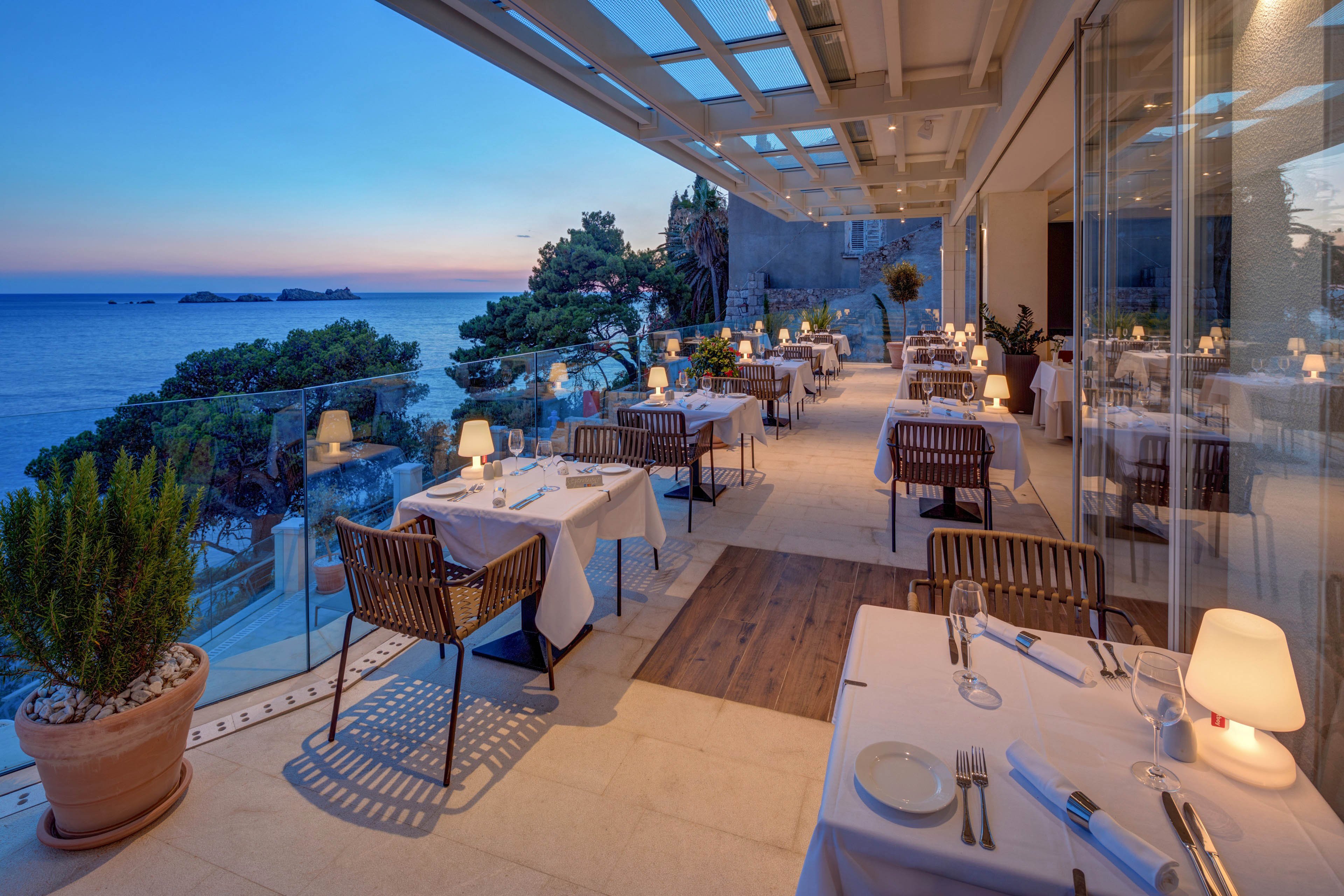 An elegant seaside restaurant terrace is set for dinner overlooking a calm ocean at sunset. White tablecloths, neatly arranged plates, and softly glowing table lamps create a refined dining atmosphere. Glass railings frame the view of the coastline and distant islands under a pastel evening sky. The scene feels tranquil and romantic, inviting guests to enjoy an outdoor meal by the water.