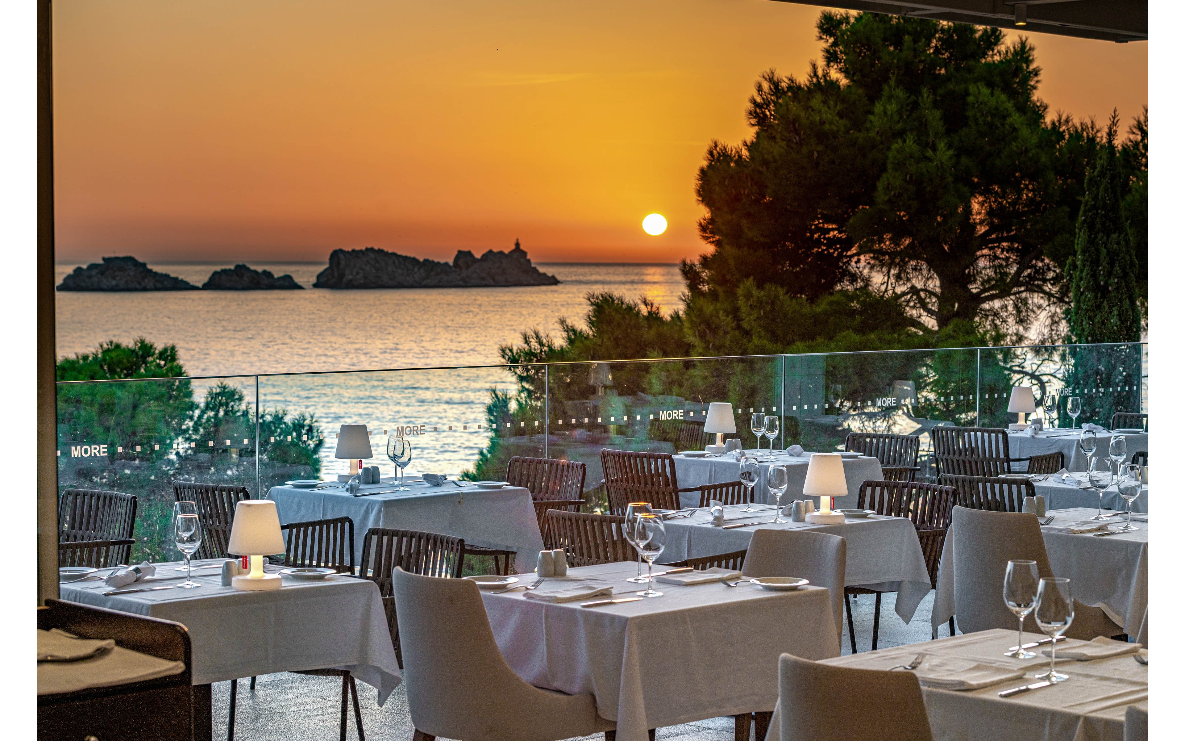 An elegant seaside restaurant terrace is set for dinner overlooking a calm ocean at sunset. White tablecloths, neatly arranged chairs, and softly glowing table lamps create a refined dining atmosphere. Glass railings frame the view of the water, rocky islets, and silhouetted trees against the orange sky. The scene suggests a tranquil, romantic evening in a coastal setting.