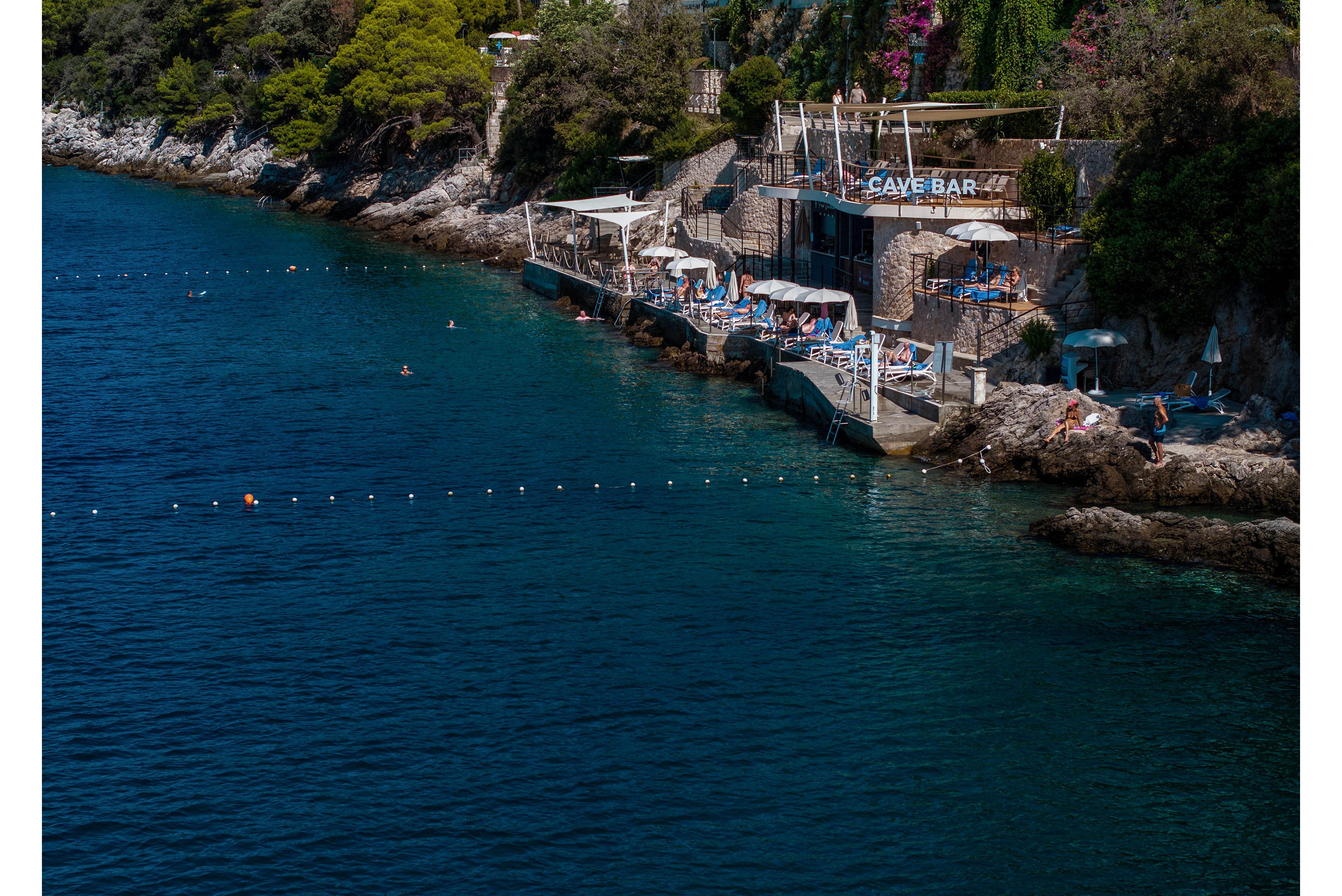 A rocky coastline features a seaside bar and sunbathing terraces built into the cliffs. People relax on lounge chairs and swim in the clear blue water near a roped-off swimming zone. The bar structure is nestled among lush greenery and trees above the stone platforms. Bright sunlight and deep turquoise sea create a vibrant, summery atmosphere.
