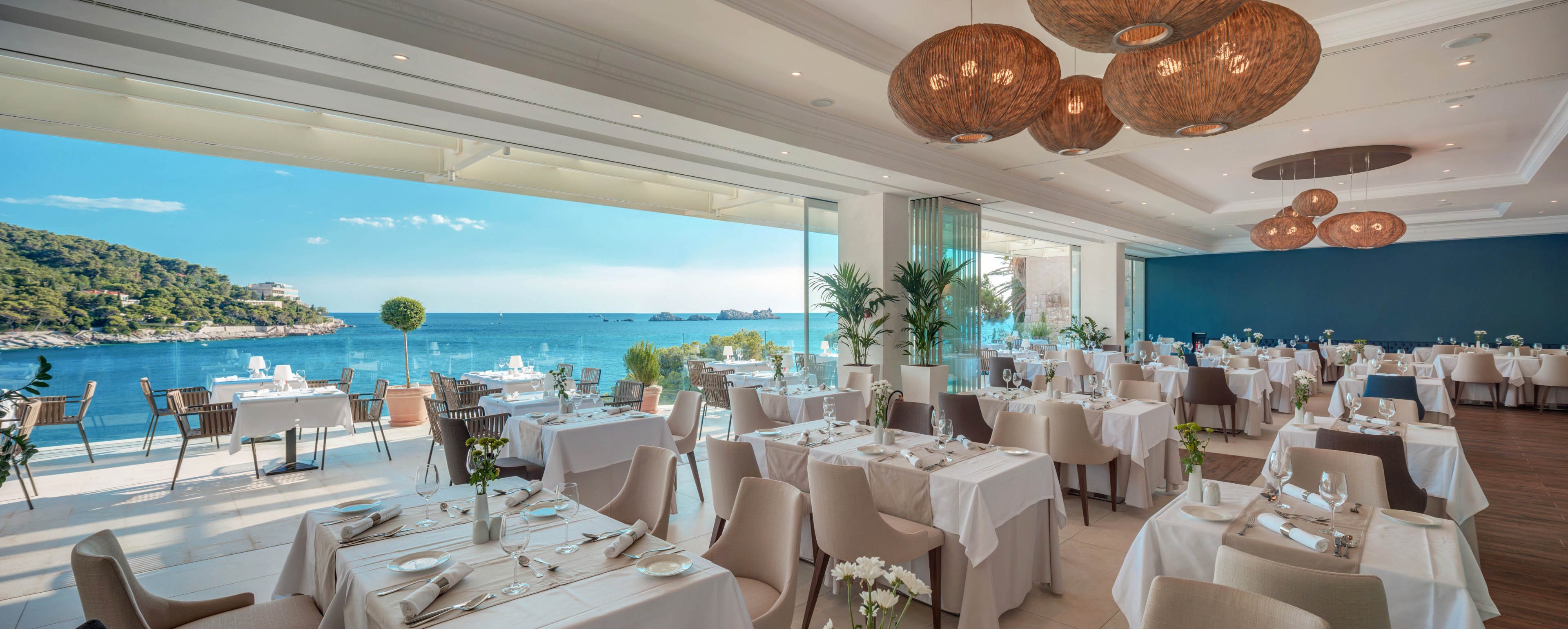 A bright, upscale restaurant dining room overlooks a stunning coastal seascape through large open windows. White tablecloths, neatly arranged place settings, and neutral-toned chairs create a refined and modern atmosphere. Natural light floods the space, highlighting decorative hanging lamps and lush indoor plants. The tranquil blue water and distant shoreline provide a serene backdrop for fine dining.