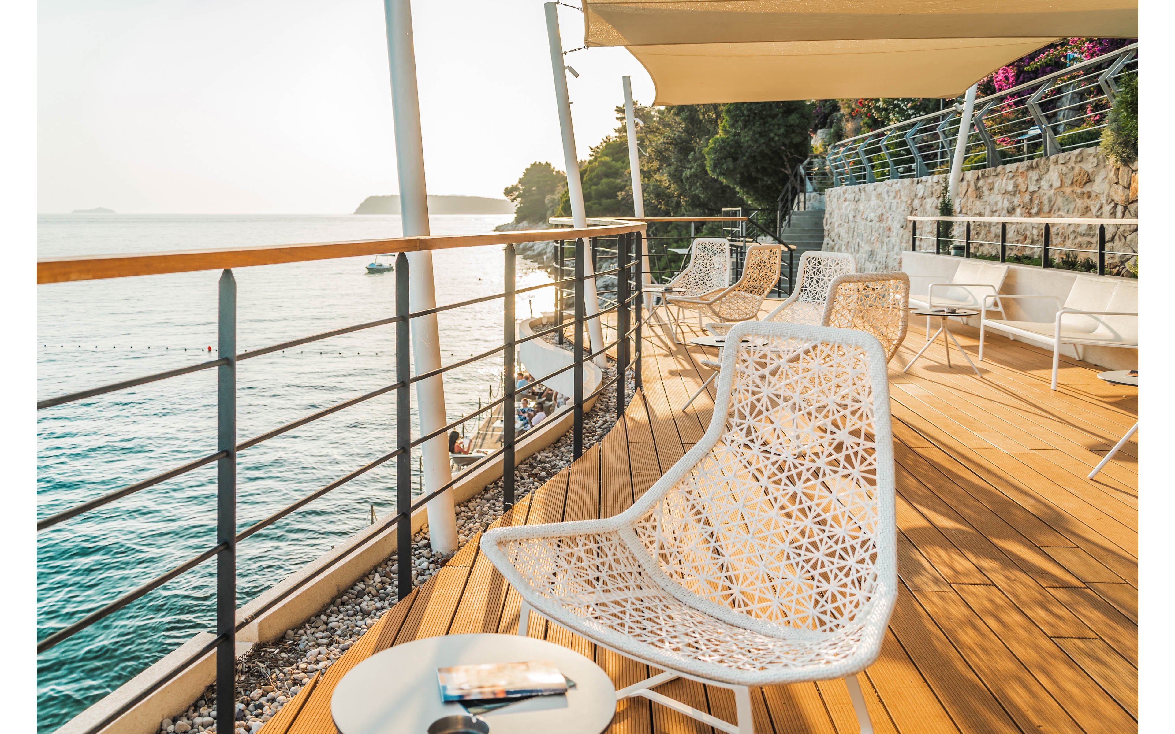 A modern seaside terrace features white woven chairs arranged on a wooden deck. The space overlooks calm blue water, separated by a metal railing running along the edge. Sunlight casts warm shadows across the deck, creating a relaxed and inviting atmosphere. In the background, additional seating and flowering plants line the stone wall, enhancing the coastal resort feel.