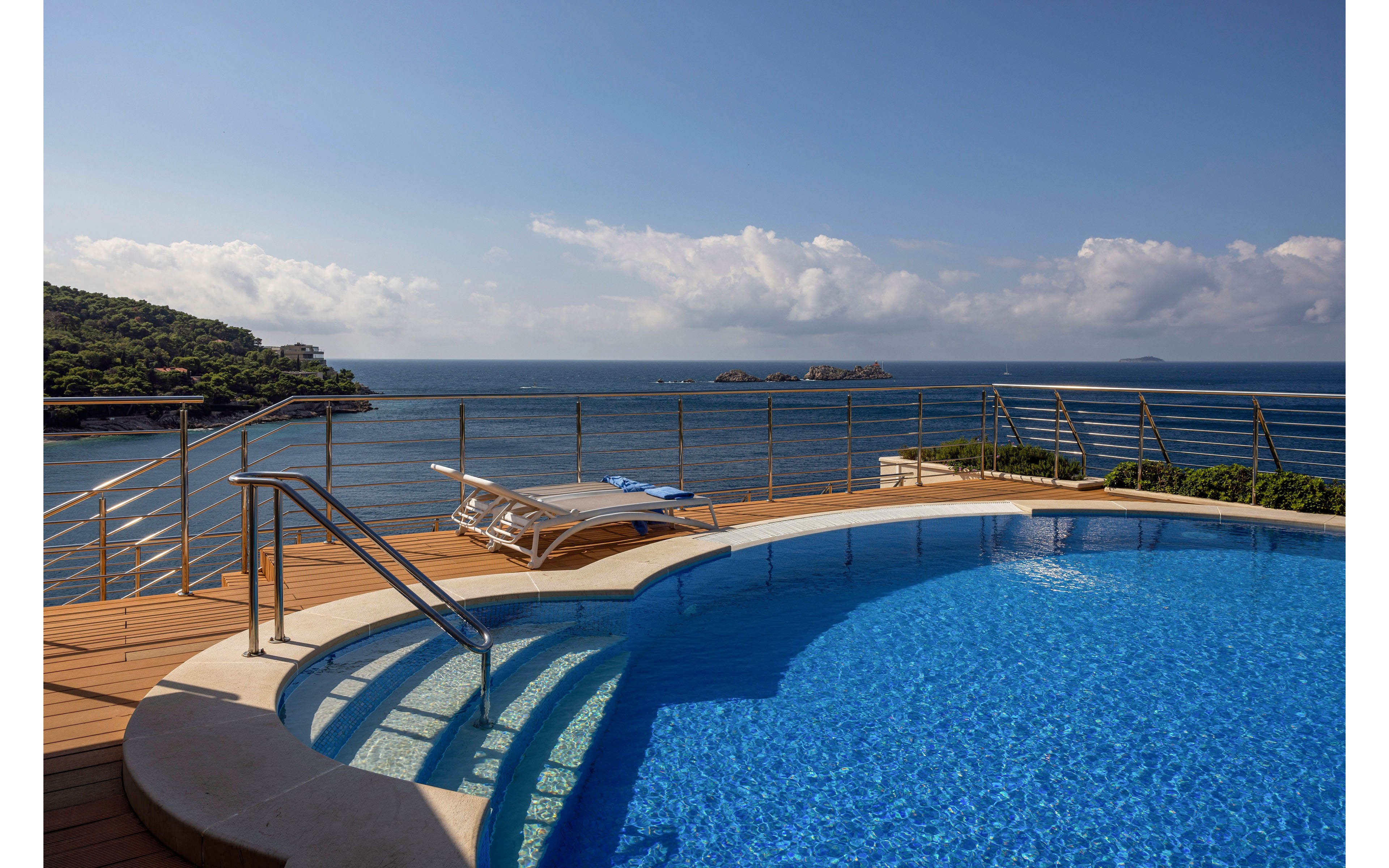 A curved infinity pool with clear blue water sits on a wooden deck overlooking a calm sea. Metal railings and a stainless steel handrail frame the pool area, leading the eye toward the horizon. A single white lounge chair rests near the edge of the deck, suggesting a quiet, relaxing retreat. Distant green hills and small rocky islets appear under a bright sky with scattered clouds.