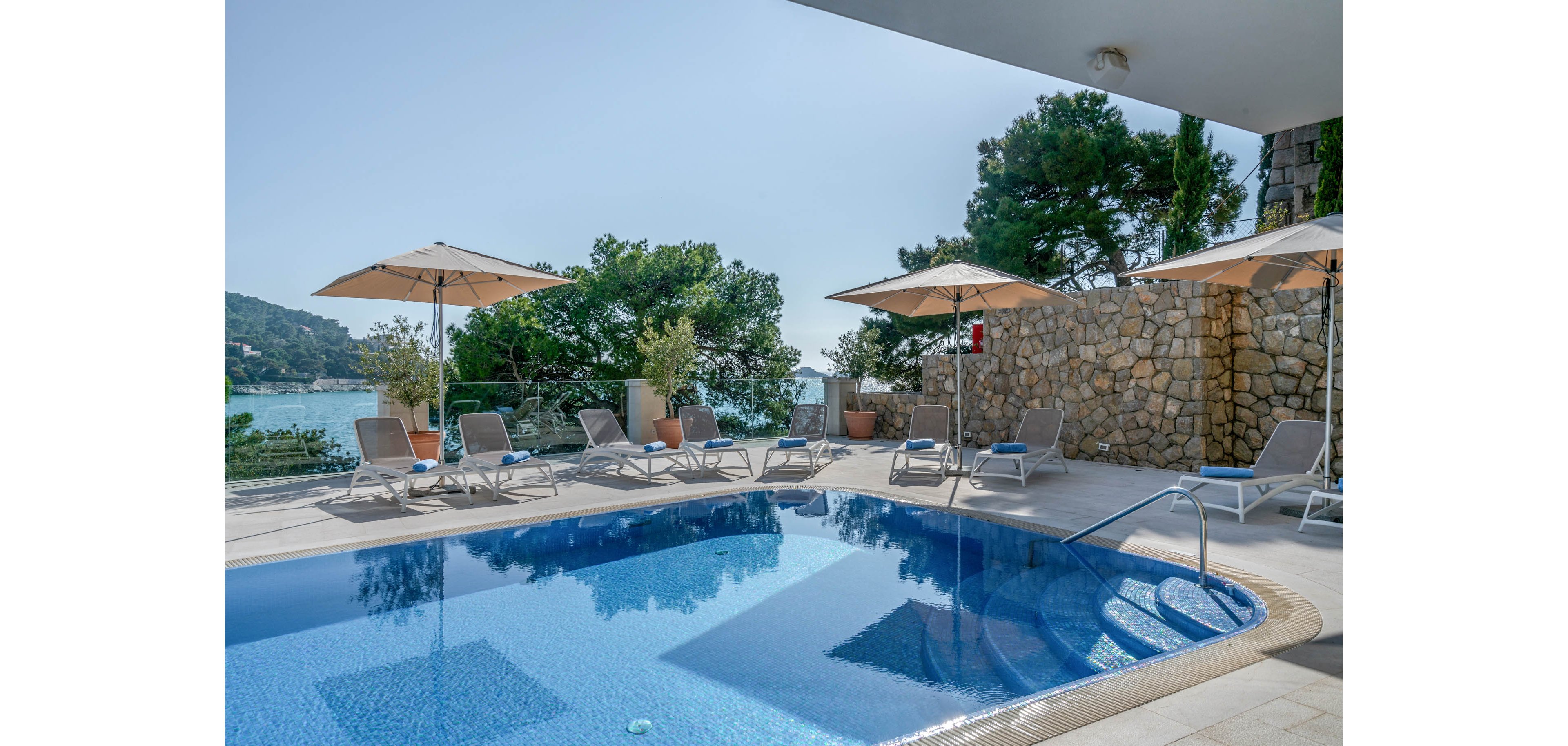 An outdoor swimming pool sits on a bright terrace overlooking a calm seaside bay. Several sun loungers and beige umbrellas are arranged around the pool, inviting relaxation. A stone wall and lush green trees frame the scene, adding a natural touch to the modern setting. Clear blue water and a cloudless sky create a tranquil, vacation-like atmosphere.