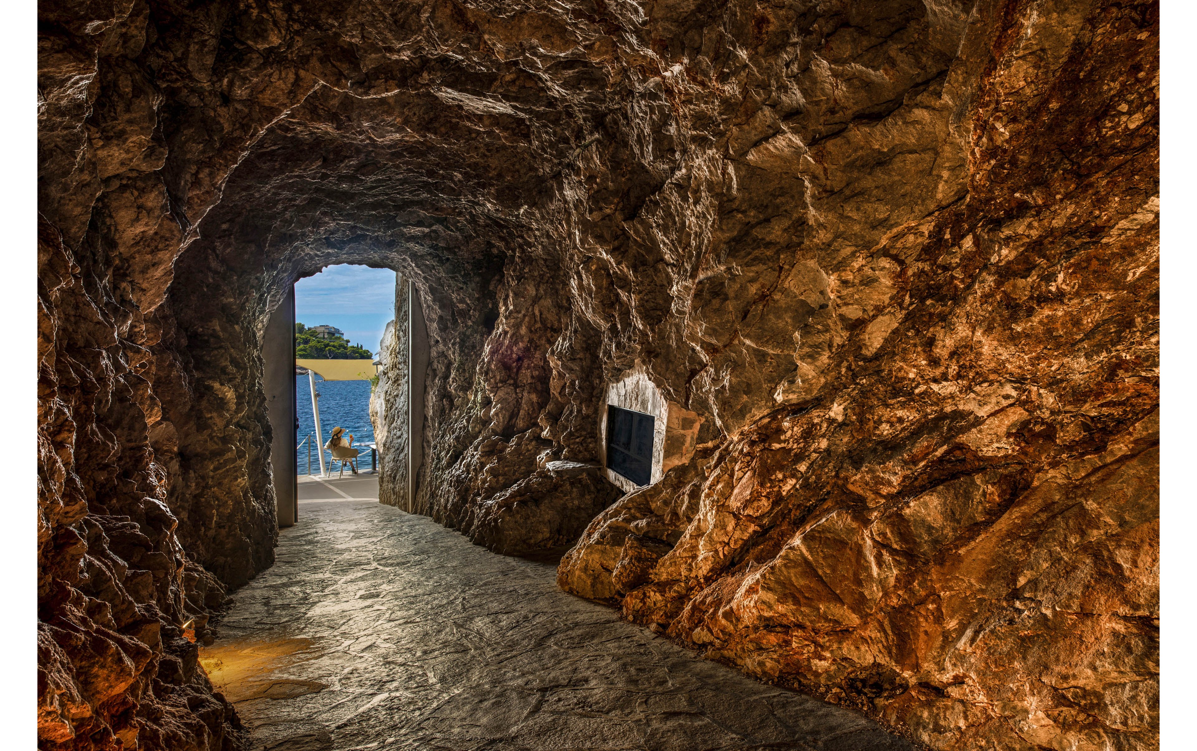A rugged stone tunnel leads toward a bright opening overlooking a calm body of water. Warm artificial lighting highlights the textured rock walls and uneven floor inside the passage. Outside, a small seating area with chairs faces the scenic waterfront and distant shoreline. The contrast between the dark cave-like interior and the sunlit coastal landscape creates a dramatic, inviting atmosphere.