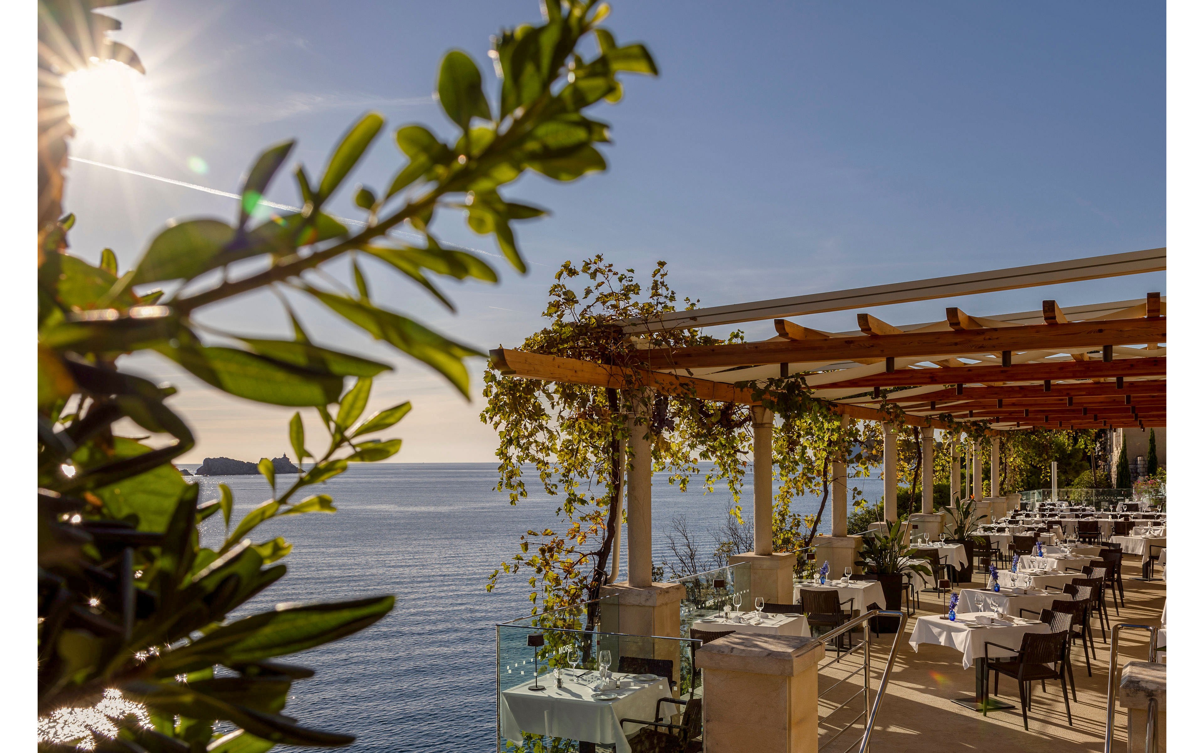 An elegant open-air restaurant terrace overlooks a calm, sunlit sea. Tables are neatly set with white tablecloths and arranged along the edge of the patio under a wooden pergola. Green vines and leafy plants frame the scene, adding a natural touch to the stone and glass architecture. The warm sunlight and clear sky create a relaxed, upscale coastal dining atmosphere.