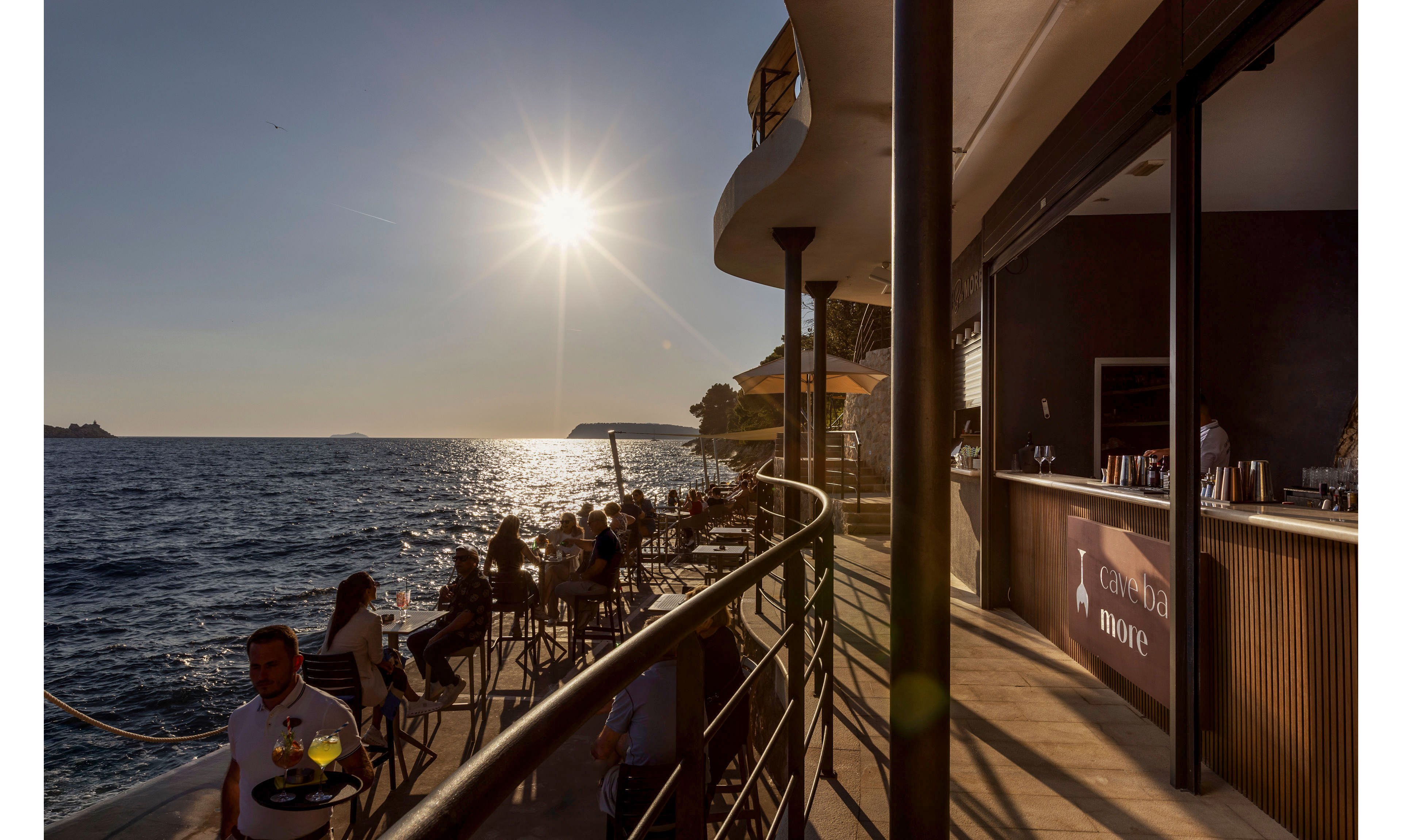 Outdoor café guests sit along a waterfront terrace as the sun sets over the sea. The scene shows people enjoying drinks and conversation beside railings overlooking calm water. Warm golden light casts long shadows and highlights the relaxed atmosphere. A bar counter is visible on the right, with text on the wall reading "Te... more".