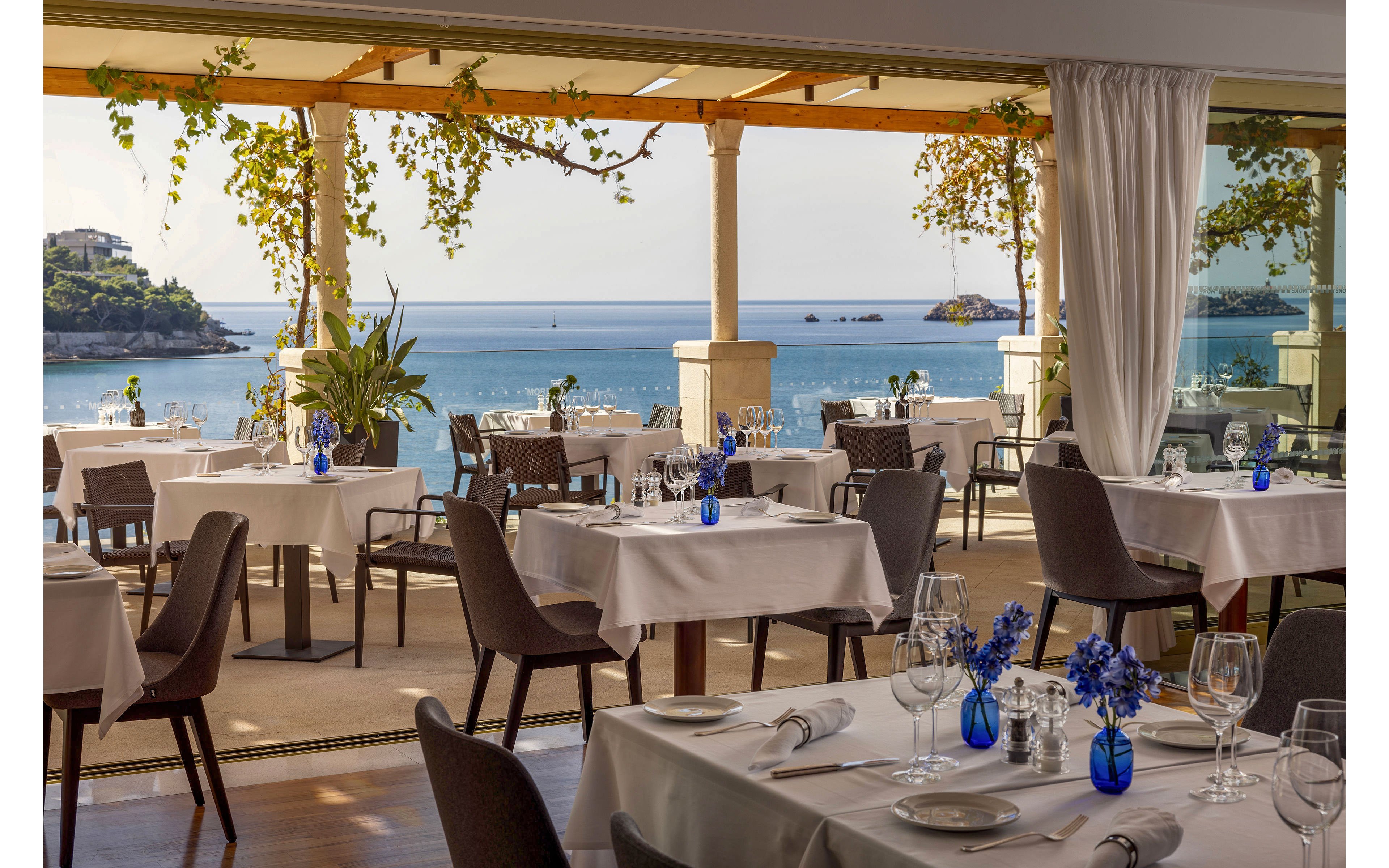An upscale restaurant dining area overlooks a calm blue sea and distant coastline. Tables are neatly set with white tablecloths, glassware, and small blue vases filled with flowers. Large windows and an open terrace allow natural light to flood the space, with greenery climbing around the pergola. The atmosphere feels serene and refined, ideal for leisurely coastal dining.