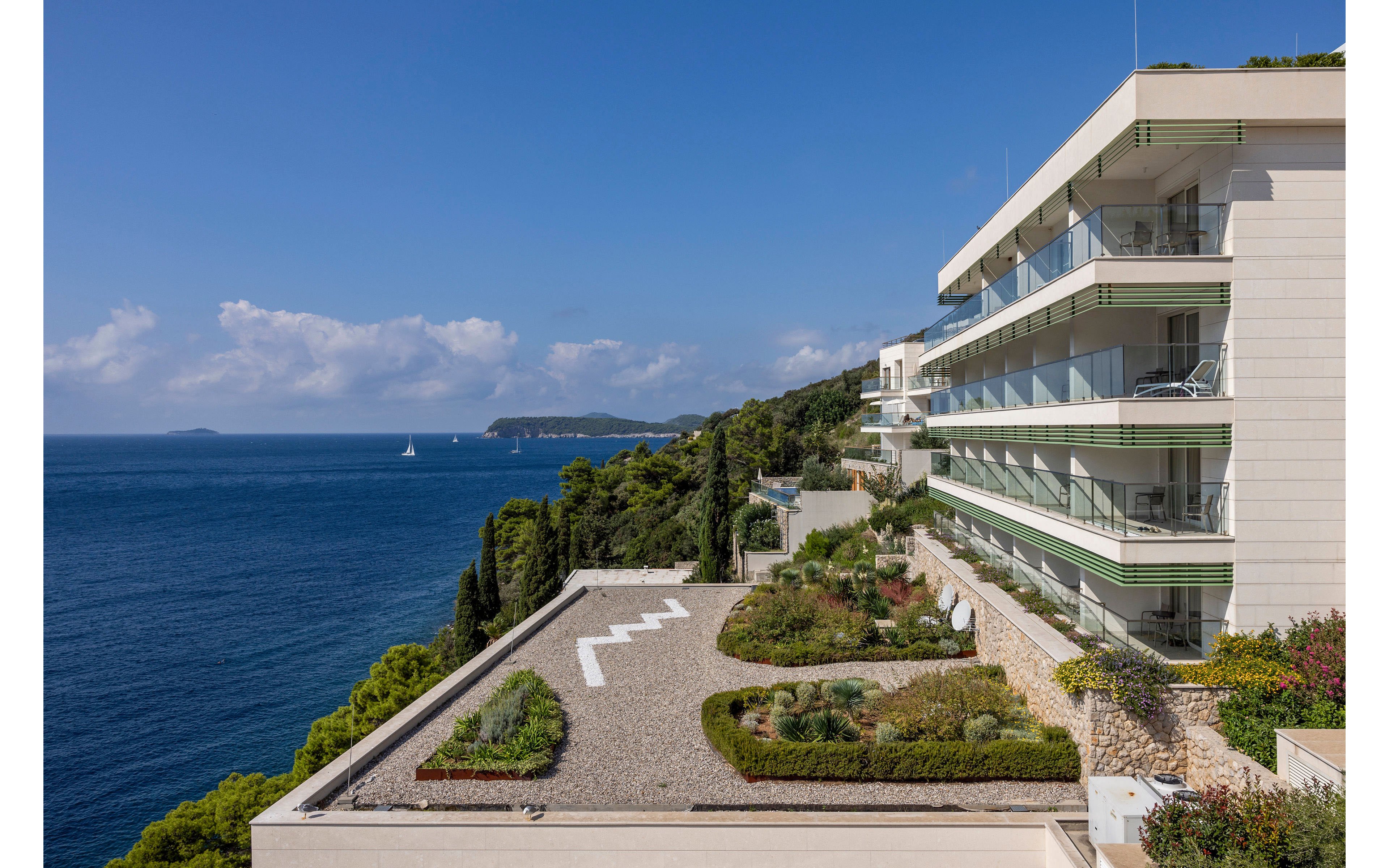 A contemporary multi-story apartment building stands on a cliffside overlooking a calm blue sea. The foreground shows a landscaped rooftop terrace with gravel paths, greenery, and geometric planting beds. Sailboats and distant islands are visible on the horizon under a clear blue sky with scattered clouds. The scene conveys a peaceful coastal lifestyle with modern architecture and lush vegetation.
