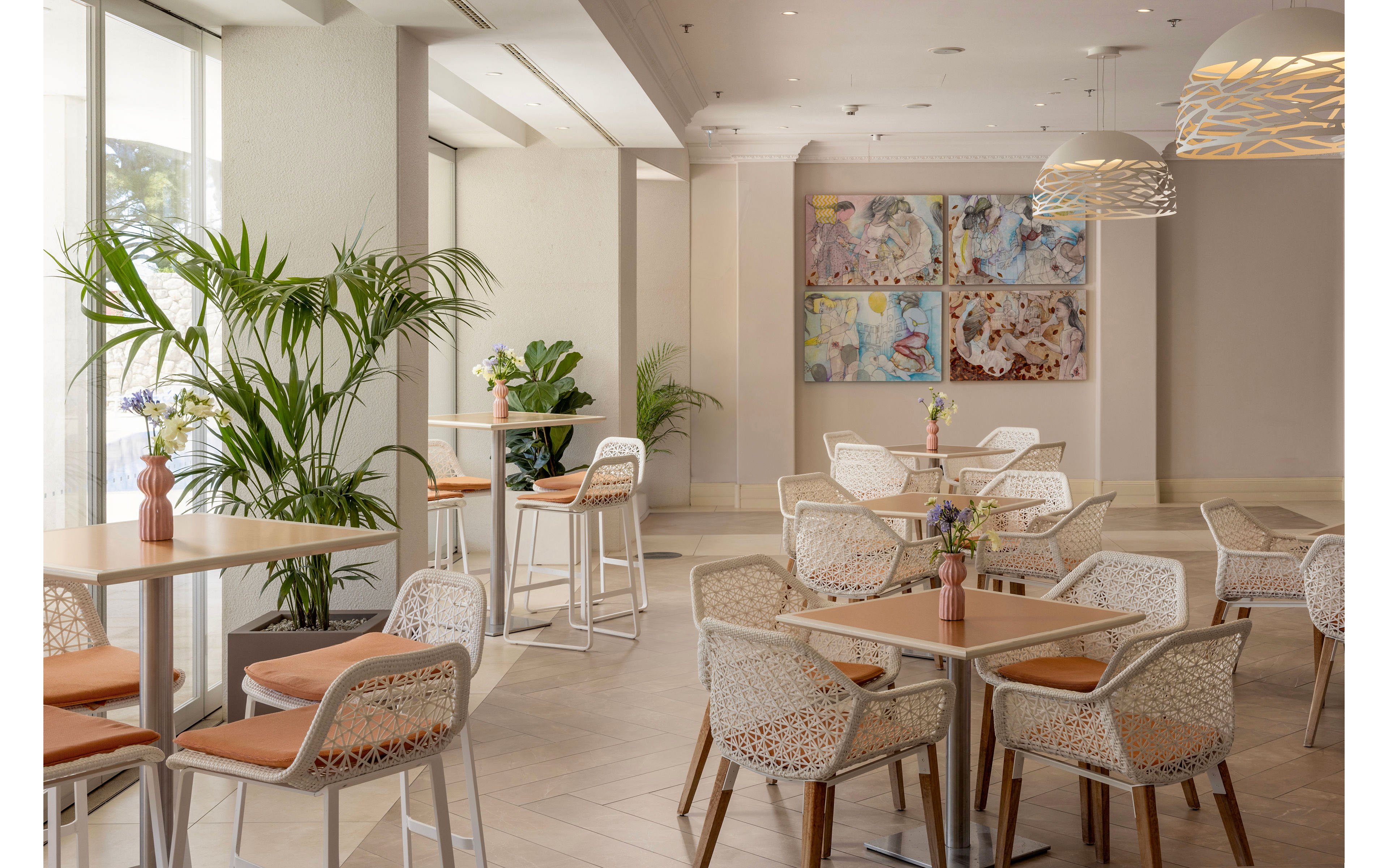 A contemporary cafe interior features light wooden tables and white woven chairs with soft orange seat cushions. Large windows on the left bring in natural daylight, illuminating potted plants and minimalist decor. Pendant lights with patterned shades hang from the ceiling, adding warmth to the neutral color palette. Colorful abstract artwork is displayed on the far wall, enhancing the relaxed and inviting atmosphere.