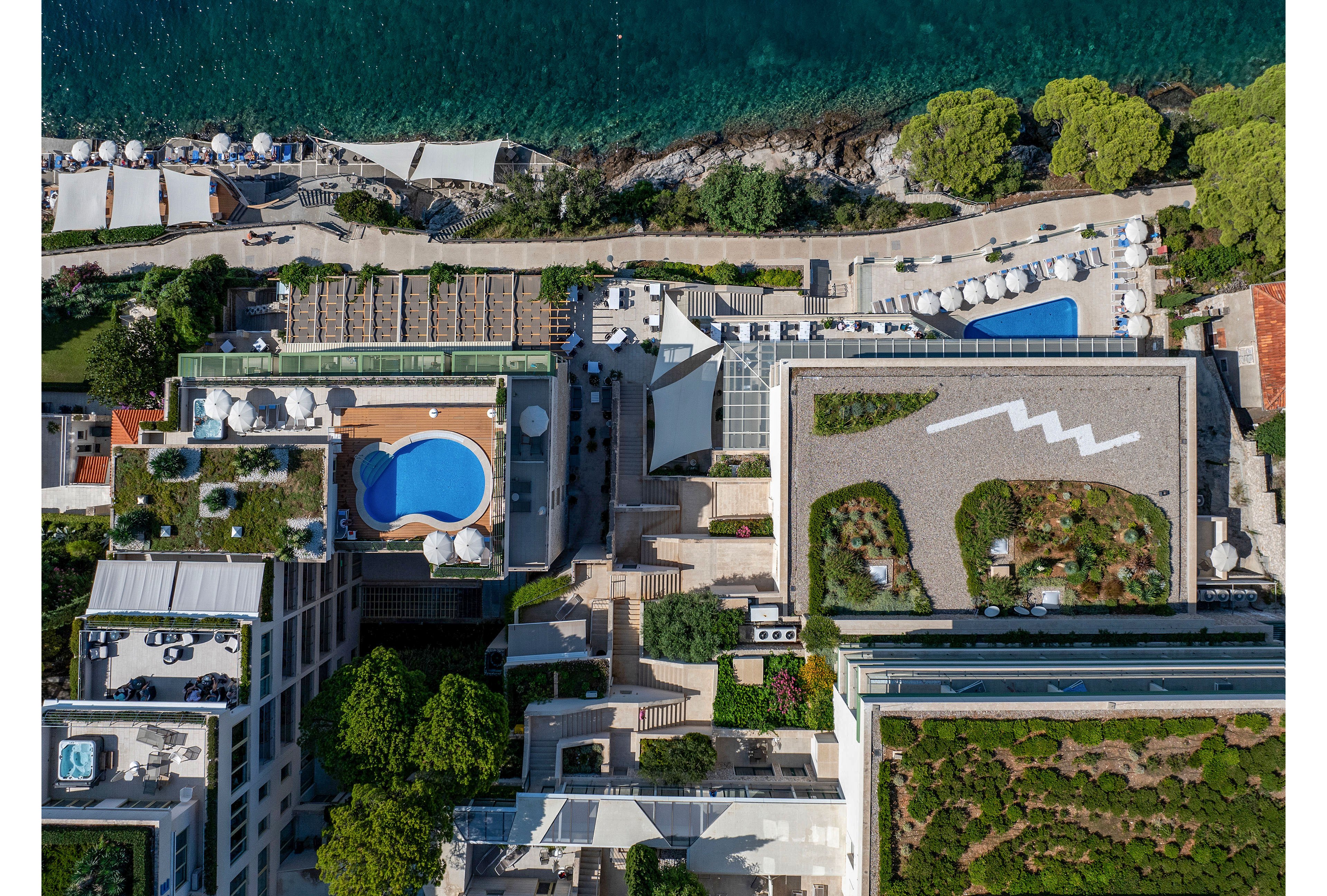 A coastal hotel complex is captured from above, showing multiple buildings and rooftop areas. A curved blue swimming pool stands out among terraces and greenery. On one rooftop, a zigzag white design contrasts with planted sections, while another features landscaped gardens. Sun loungers and umbrellas line the waterfront beside the clear blue sea, emphasizing a resort setting.