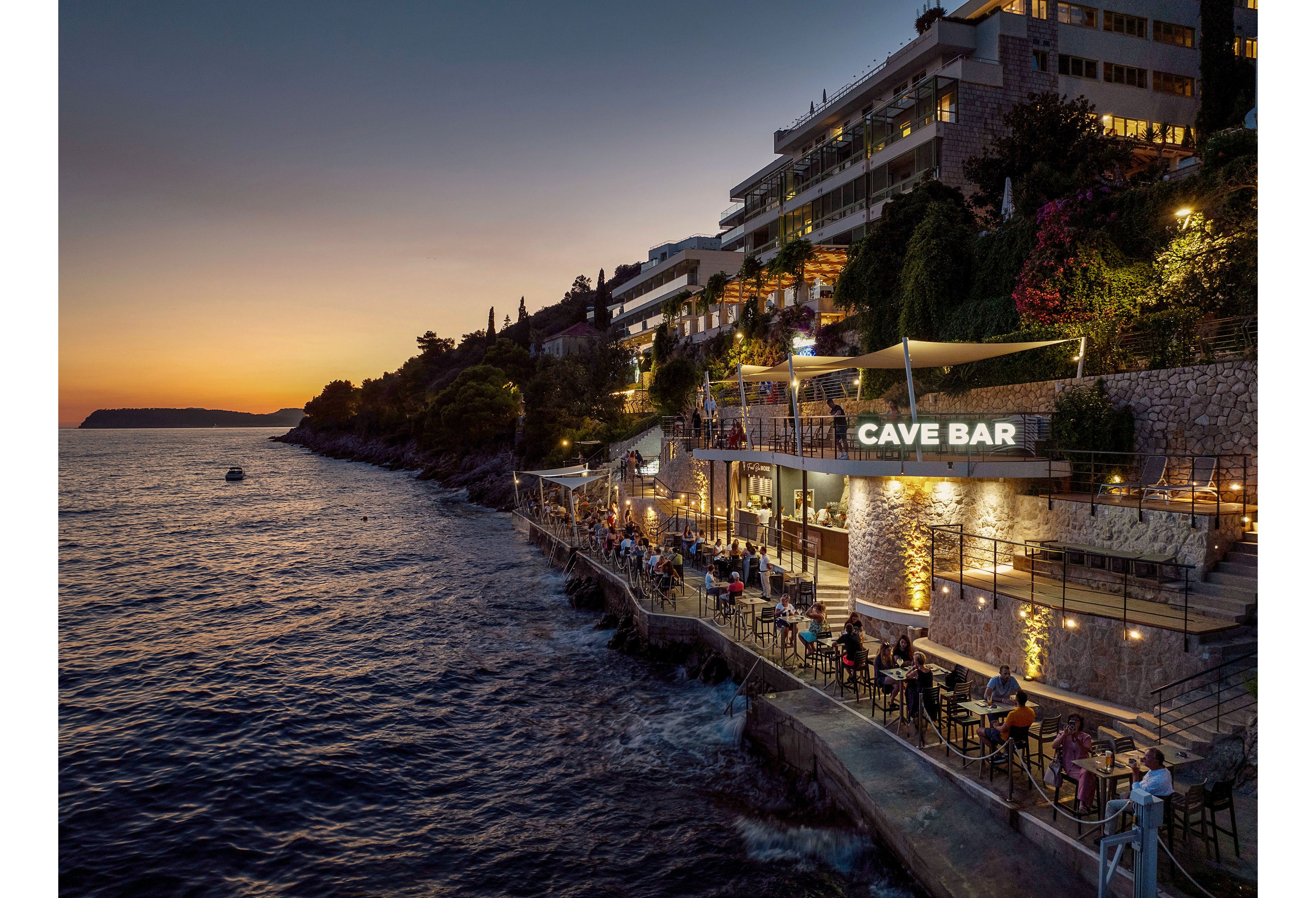A lively cave bar terrace overlooks the sea at sunset, with people dining and drinking along the waterfront. The bar is built into a rocky cliff below a row of modern hillside buildings and hotels. Warm artificial lights contrast with the deep blue water and fading orange sky, creating a cozy evening atmosphere. The scene captures coastal nightlife with outdoor seating, railings, and pathways hugging the shoreline.