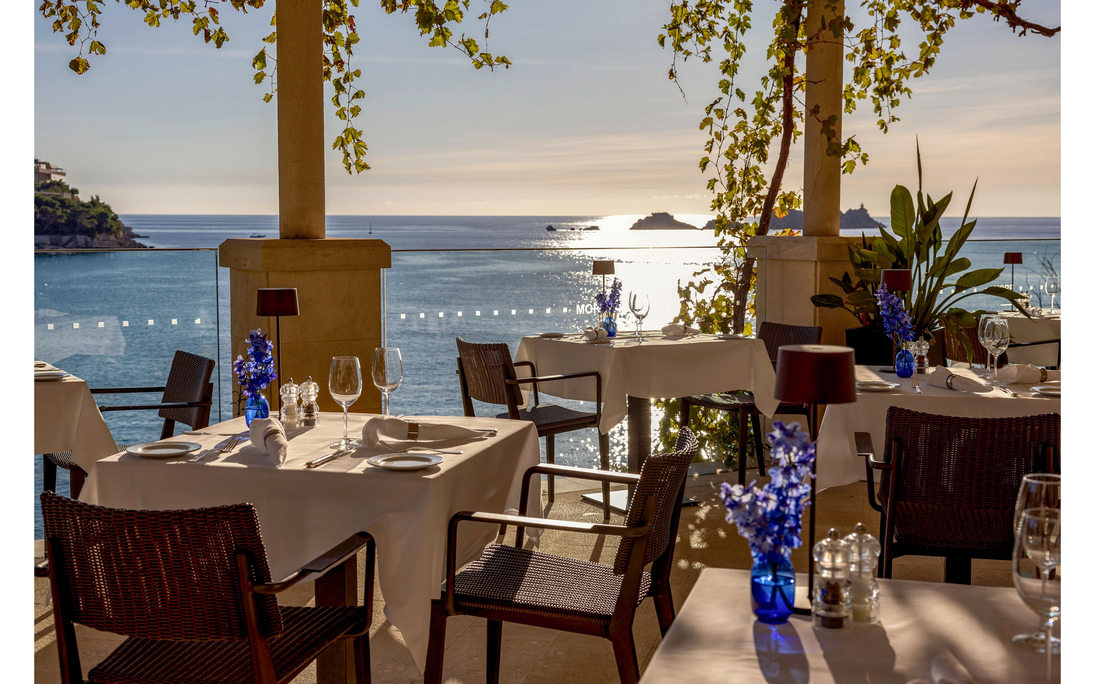An upscale outdoor restaurant terrace overlooks a calm sea at sunset. Tables are set with white tablecloths, glassware, and cutlery, creating a fine dining atmosphere. Blue vases, plants, and warm-toned lamps add decorative accents to the scene. Sunlight reflects off the water, with distant islands and coastal buildings visible in the background.