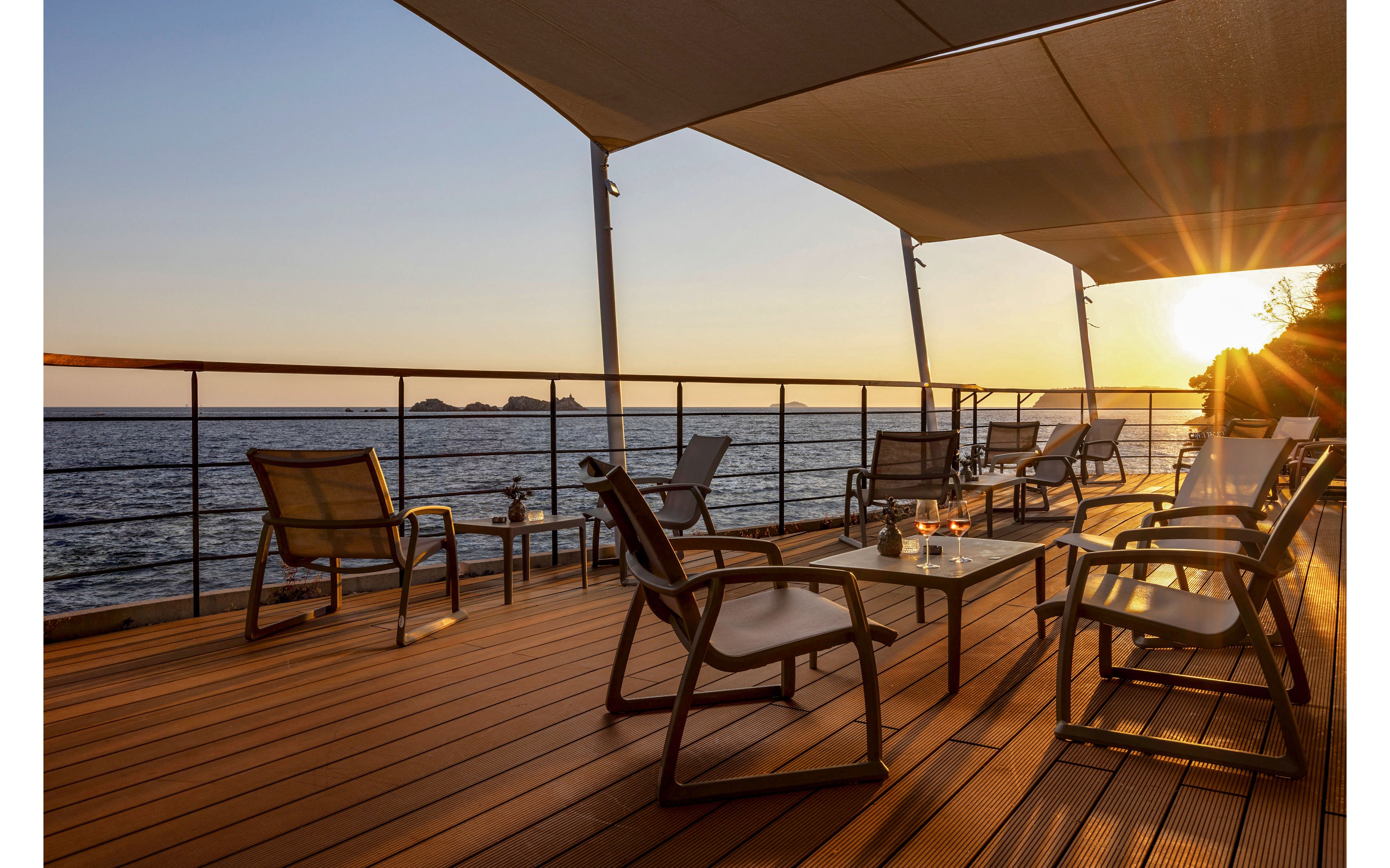 An open-air terrace overlooks a calm sea at sunset, with warm sunlight streaming across the wooden deck. Several lounge chairs and small tables are arranged facing the water, inviting relaxation and quiet conversation. The sun is low on the horizon, casting golden reflections on the ocean and creating long shadows across the seating area. A simple railing and overhead shade structures frame the view of distant islands and the glowing sky.