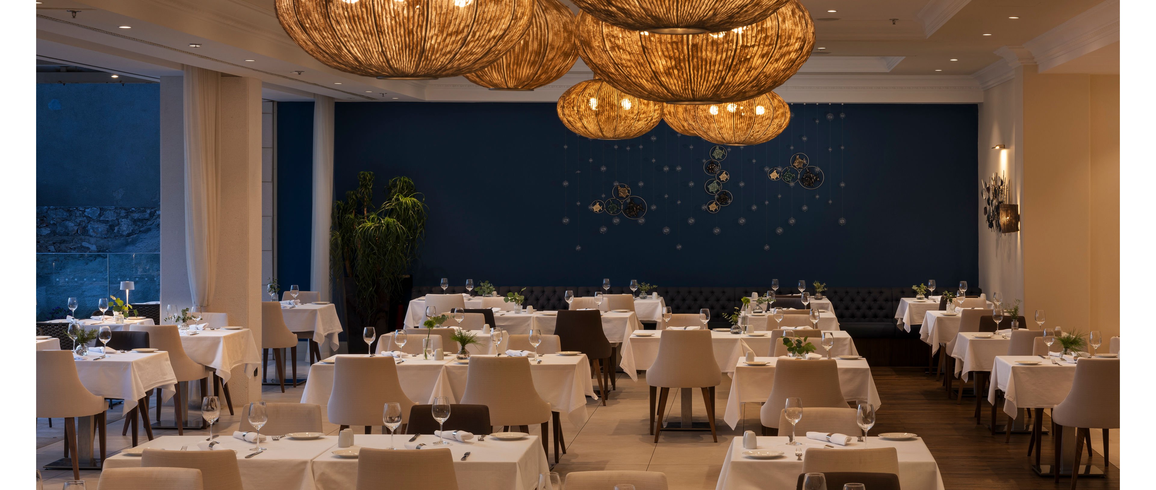 Upscale restaurant dining room with neatly arranged tables covered in white tablecloths is shown in a calm evening setting. Contemporary wicker pendant lights hang from the ceiling, casting a warm glow over the space. A deep blue accent wall with decorative hanging elements adds visual interest behind the seating area. The room appears prepared for guests, with glassware, cutlery, and chairs carefully positioned around each table.