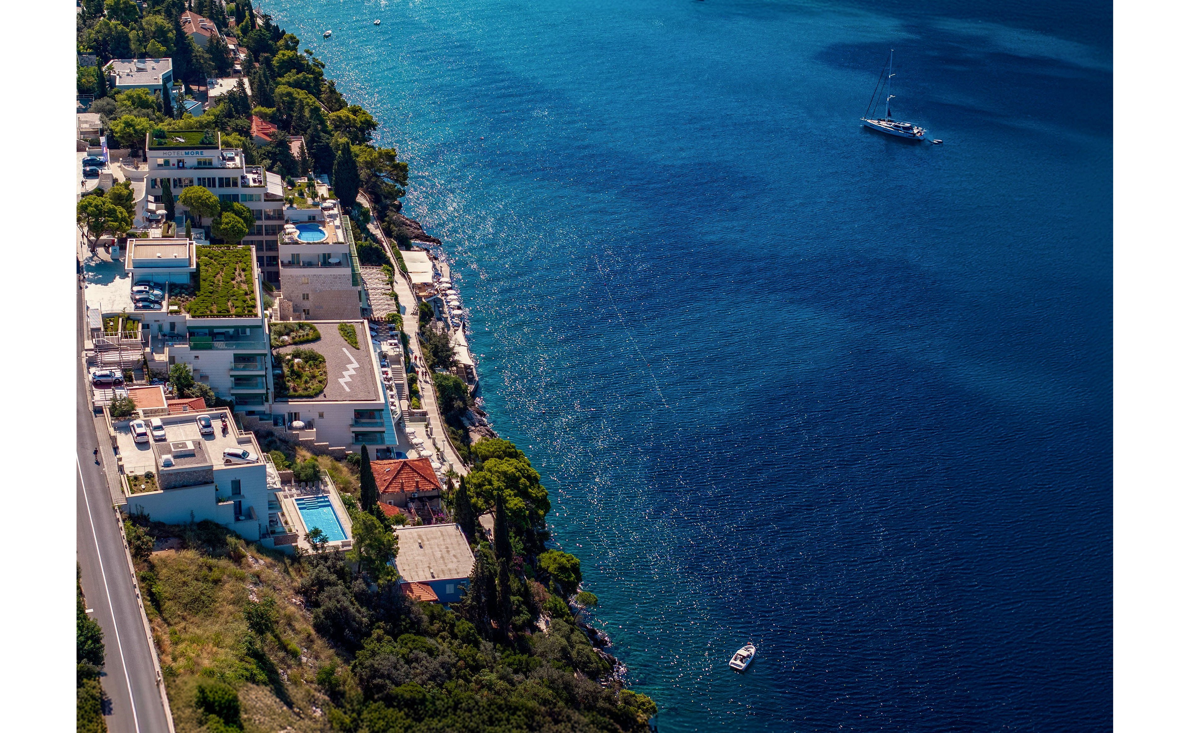 An aerial view captures modern coastal villas built along a steep shoreline. A winding road runs parallel to the water, with houses featuring terraces, pools, and lush greenery. The calm, deep blue sea dominates the right side of the image, where a sailboat and a small motorboat float peacefully. The scene conveys a serene Mediterranean-style seaside lifestyle with clear weather and vibrant colors.