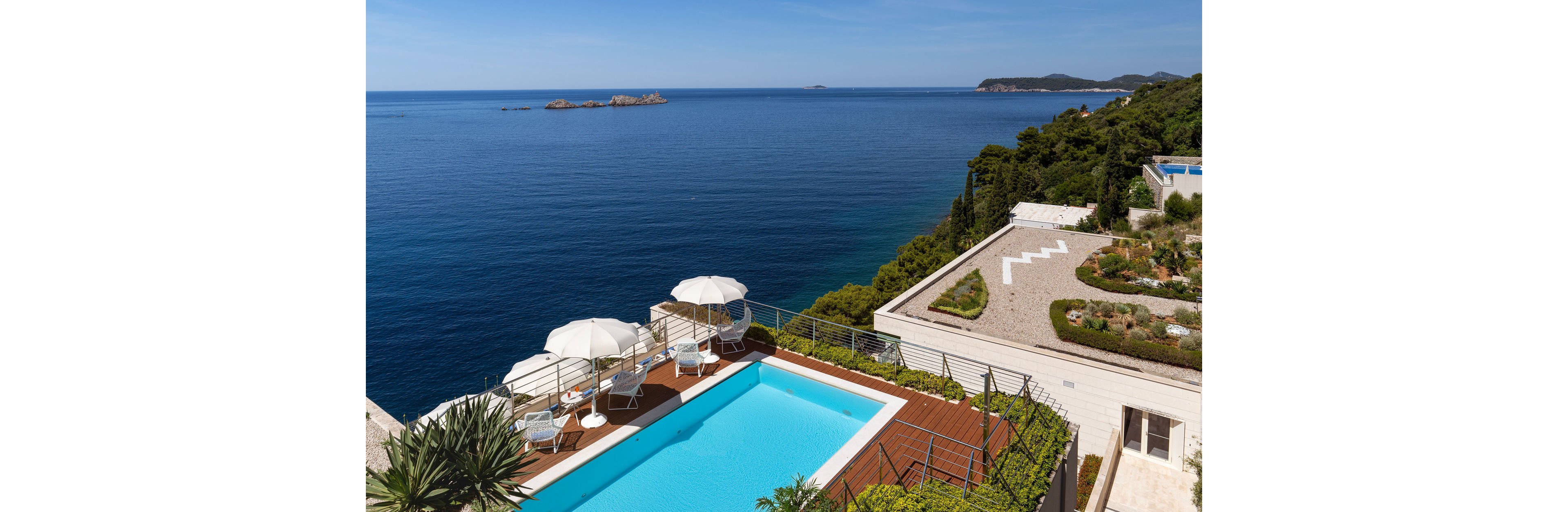 A bright turquoise swimming pool sits on a clifftop terrace above a deep blue sea. White sun loungers and umbrellas surround the pool, creating a relaxing vacation atmosphere. The coastline is lined with green trees and landscaped paths, with small islands visible in the distance. Clear skies and sunlight enhance the serene, luxurious seaside setting.