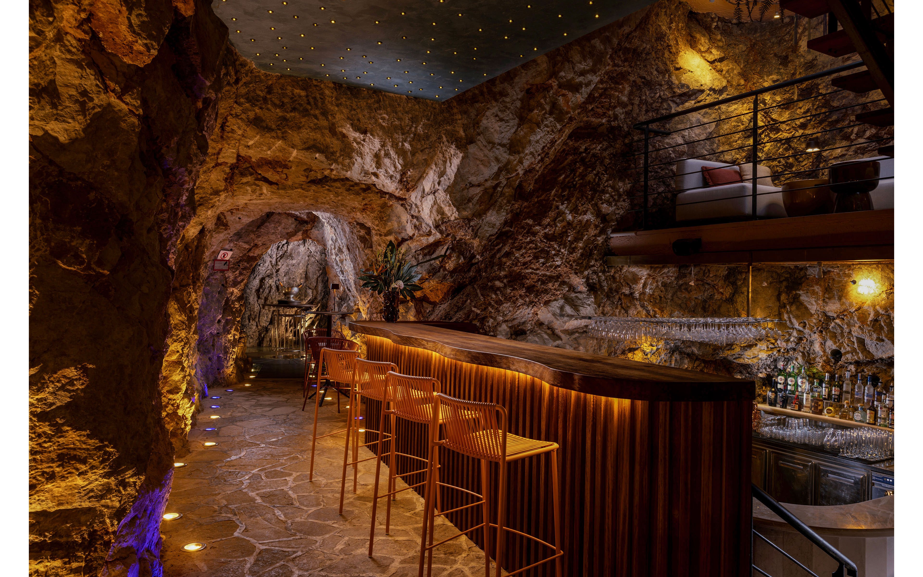 A stylish bar is built into a rocky cave interior with a long wooden counter and modern bar stools. Warm accent lighting traces the contours of the bar and floor, while tiny ceiling lights create a starry night effect above. Bottles and glassware are arranged along the back wall, and an upper mezzanine seating area overlooks the bar. The overall scene feels intimate and sophisticated, blending natural stone textures with contemporary design.