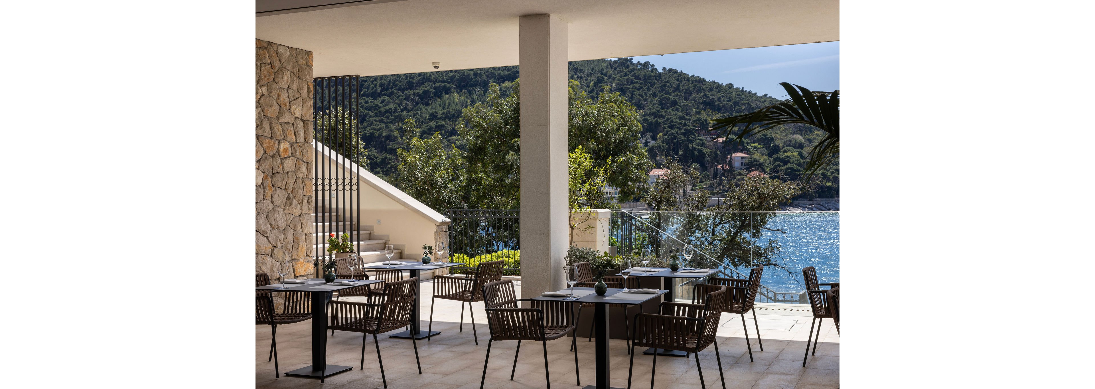An open-air restaurant terrace features neatly arranged tables and dark wooden chairs under a shaded roof. The setting overlooks a calm blue sea framed by lush green hills and scattered coastal houses. Stone walls, staircases, and metal railings add architectural interest to the space. Small table decorations and natural light create a relaxed, inviting atmosphere for waterfront dining.