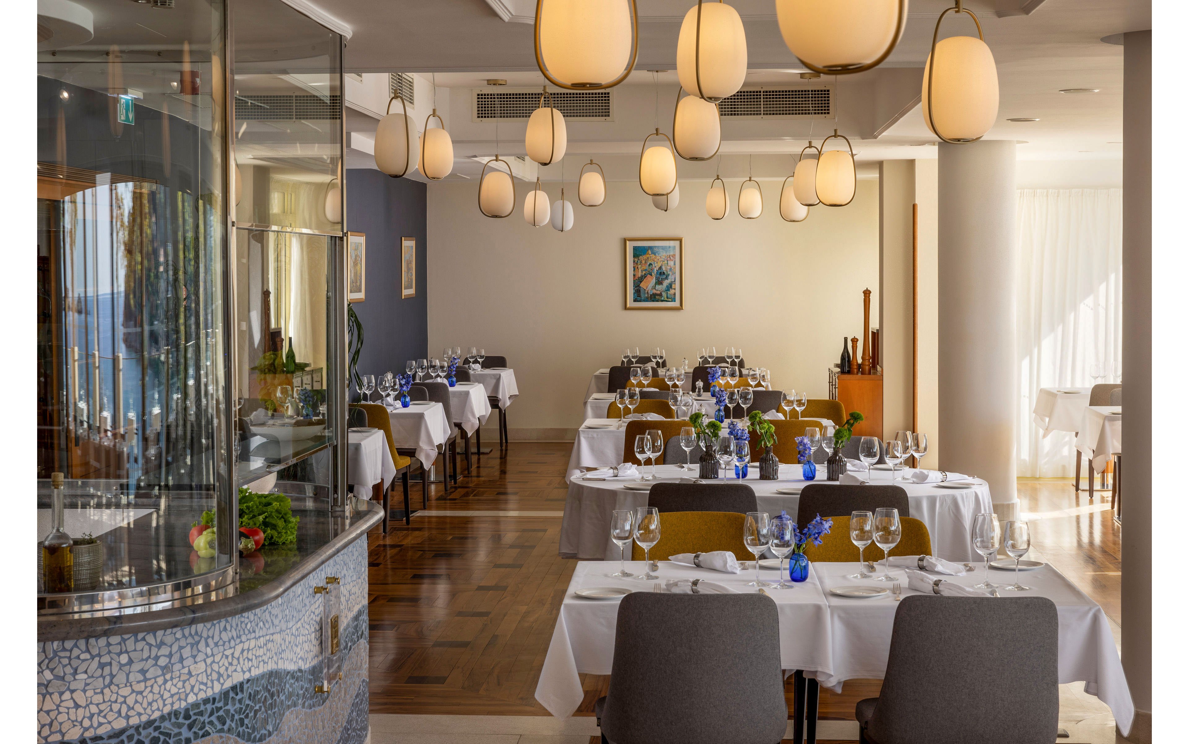 A refined restaurant dining room is set with white tablecloths and neatly arranged glassware. Tables are decorated with small blue vases and flowers, creating a fresh and inviting atmosphere. Warm pendant lights hang from the ceiling, illuminating the polished wooden floor and contemporary furnishings. Large windows with sheer curtains allow natural daylight to fill the space, highlighting the modern decor and artwork on the wall.