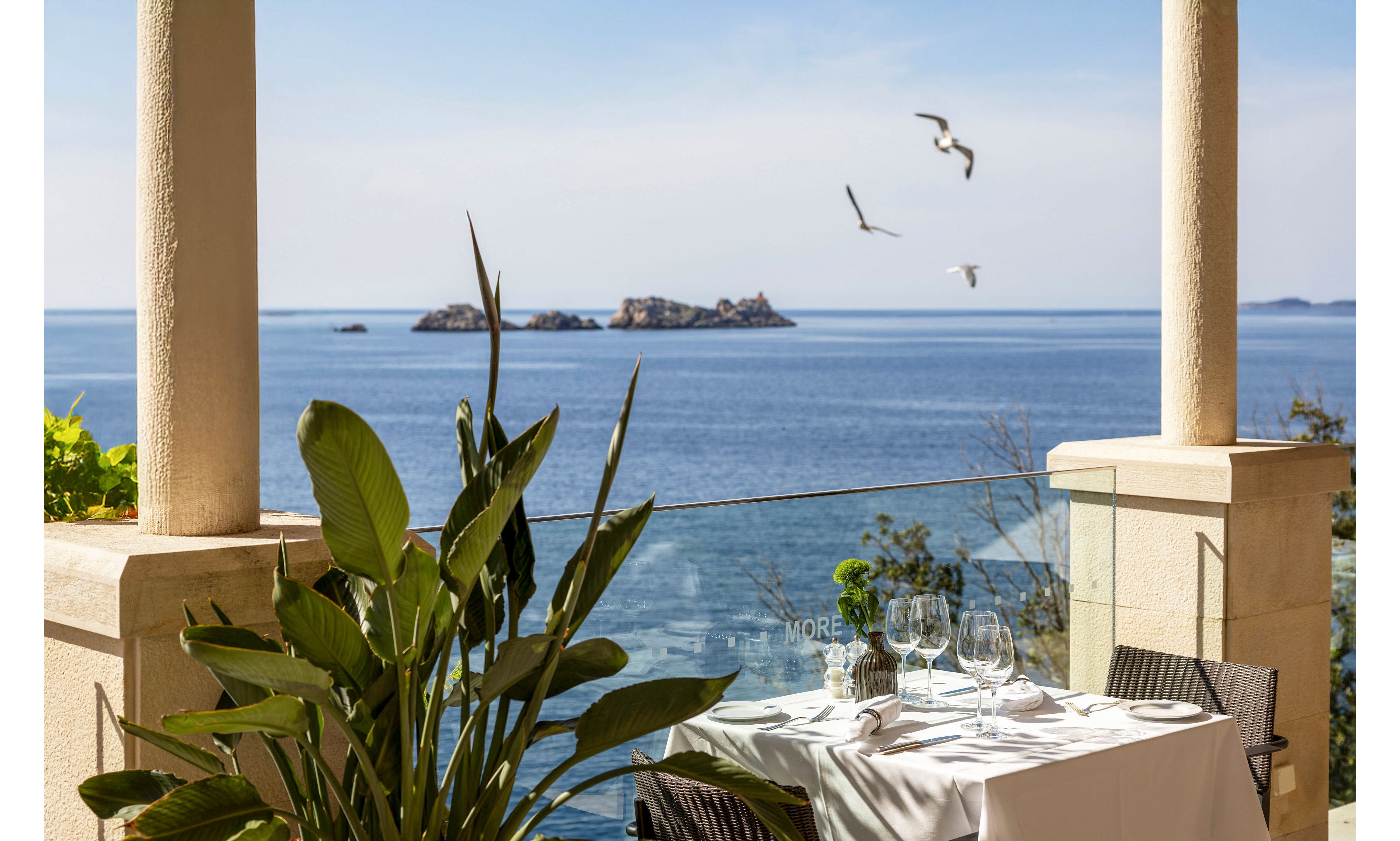 An elegant outdoor restaurant table is set with white linens, glassware, and cutlery on a terrace overlooking a calm blue sea. Lush green plants frame the foreground, while stone columns and a glass railing define the space. In the distance, rocky islets rise from the water and several seagulls fly across the clear sky. The scene suggests a peaceful coastal dining experience in warm, sunny weather.