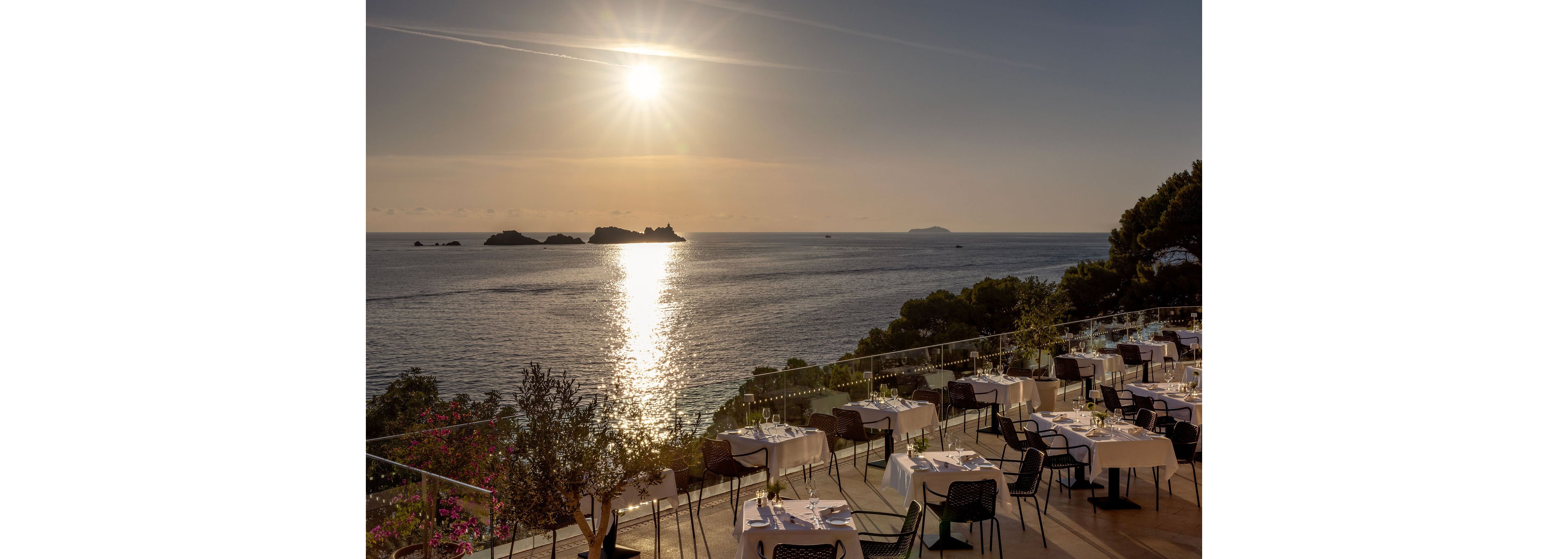 An elegant open-air restaurant terrace overlooks a calm sea at sunset. White-clothed tables and neatly arranged chairs line the stepped patio, ready for diners. The sun hangs low over the horizon, casting a bright reflection across the water toward distant rocky islets. Lush greenery and flowering plants frame the scene, enhancing the tranquil coastal atmosphere.