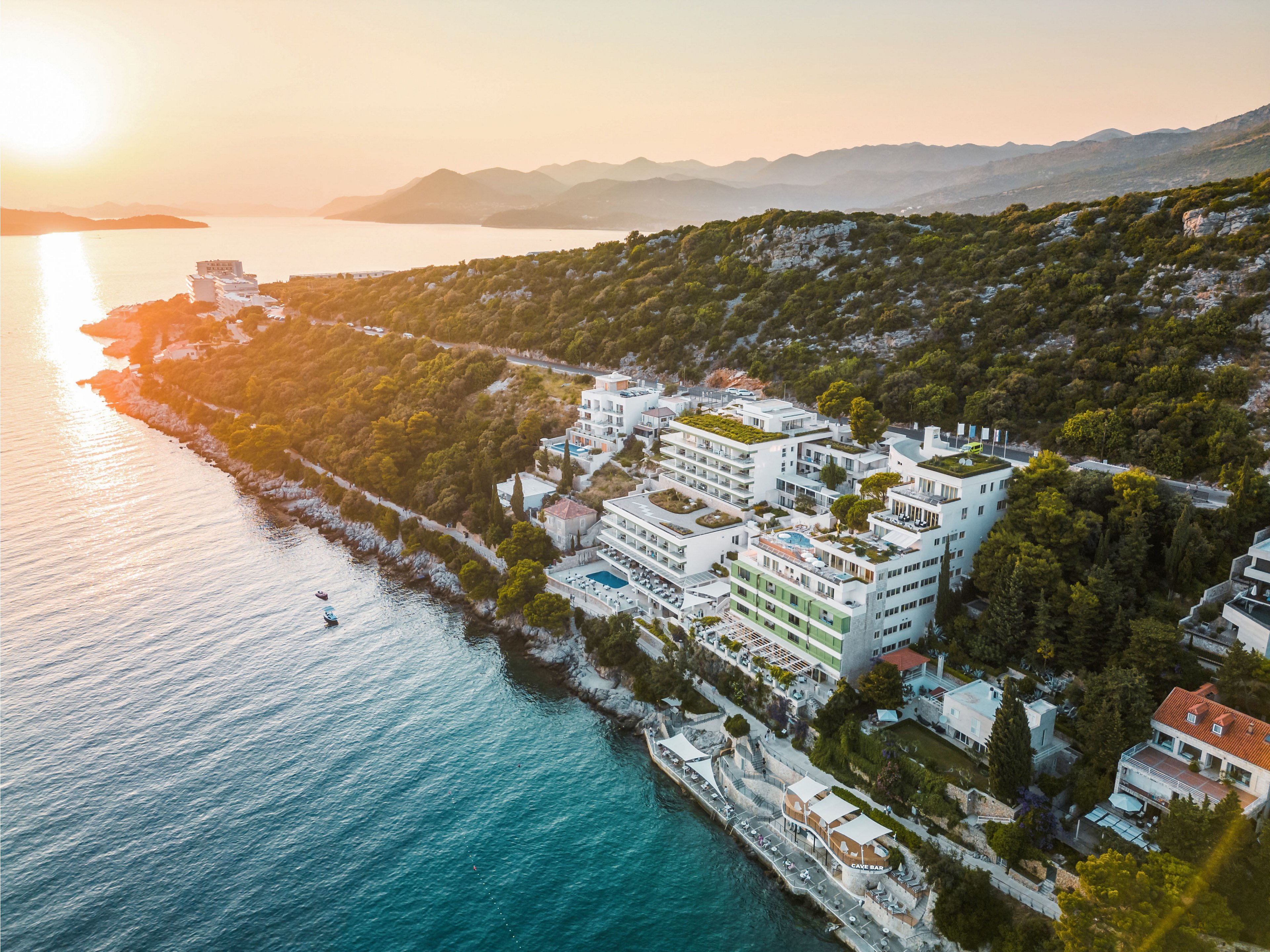A large seaside resort complex stretches along a rocky coastline beside calm blue water. The scene is captured from above, showing terraced buildings, pools, and greenery nestled against a forested hillside. Warm sunlight from the setting sun bathes the landscape in golden tones, highlighting the contrast between the sea and the lush vegetation. Distant mountains and islands fade into the horizon, adding depth and a tranquil vacation atmosphere.