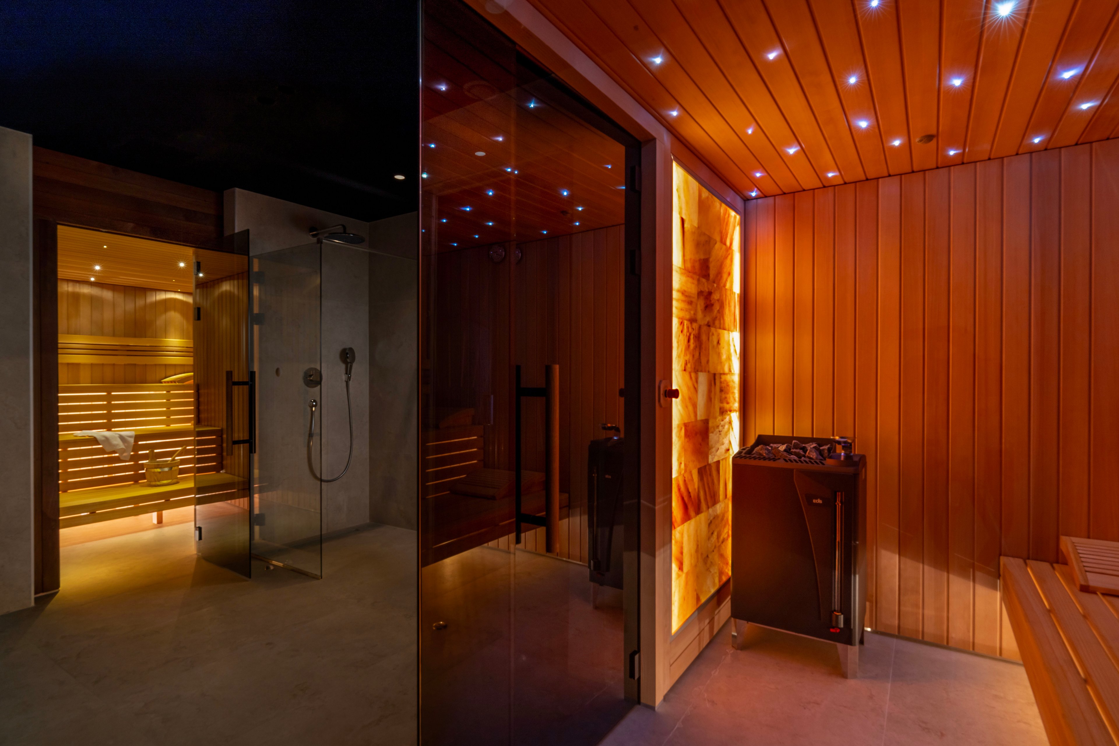 A contemporary sauna room features sleek wooden paneling and glass doors, creating a warm and inviting wellness space. Soft amber lighting glows from within the sauna, contrasting with the darker adjacent area that includes a shower. A heater unit stands near the entrance, while the ceiling is dotted with small blue lights that resemble stars. The overall scene conveys a high-end spa environment focused on relaxation and comfort.