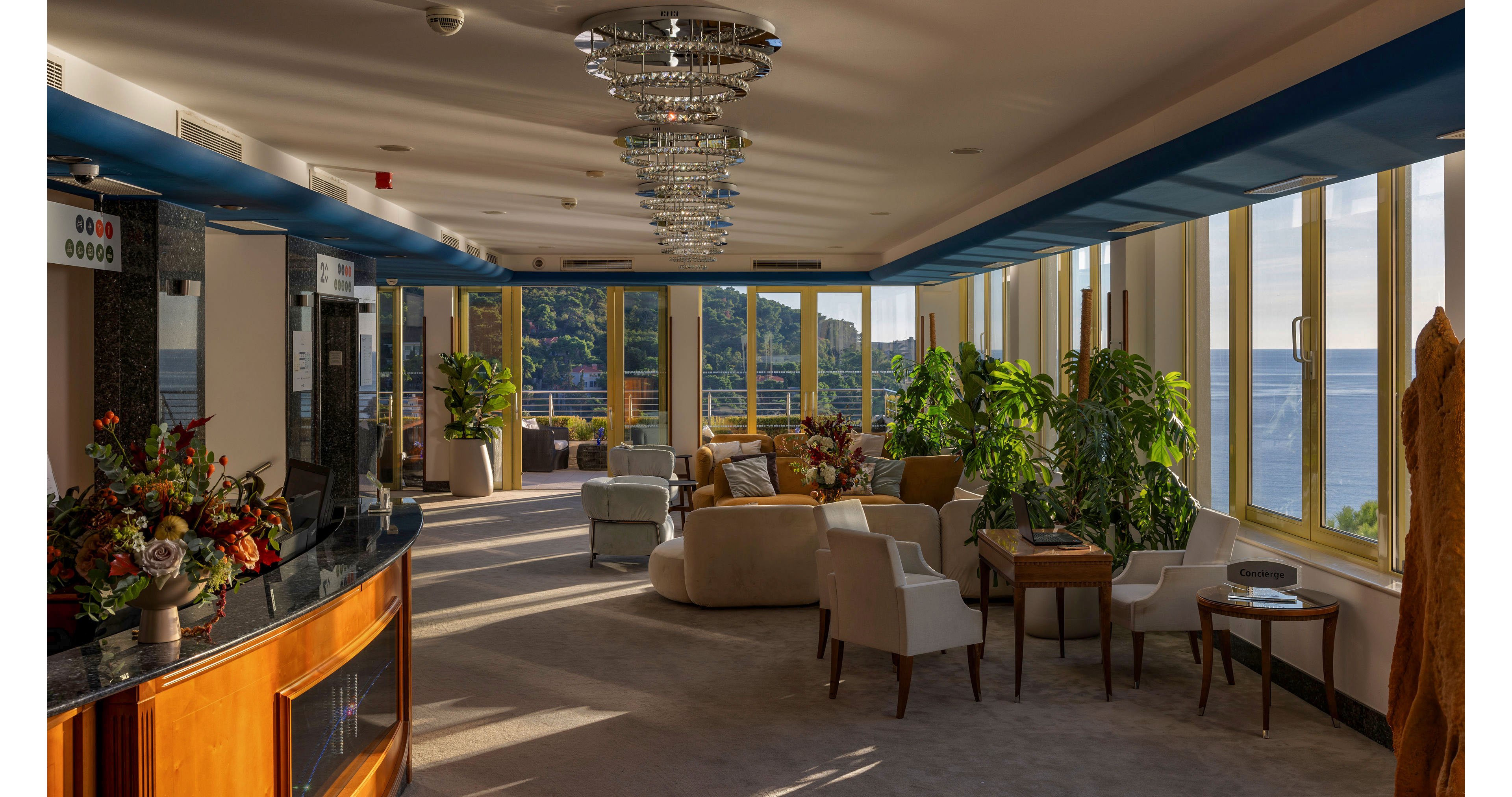 A spacious lounge area is filled with soft armchairs and sofas arranged for relaxed seating. Large windows line the room, revealing a scenic view of the sea and green hills in warm afternoon light. Decorative plants, floral arrangements, and crystal chandeliers add an elegant touch to the interior. The overall atmosphere is calm and inviting, blending modern comfort with a coastal setting.