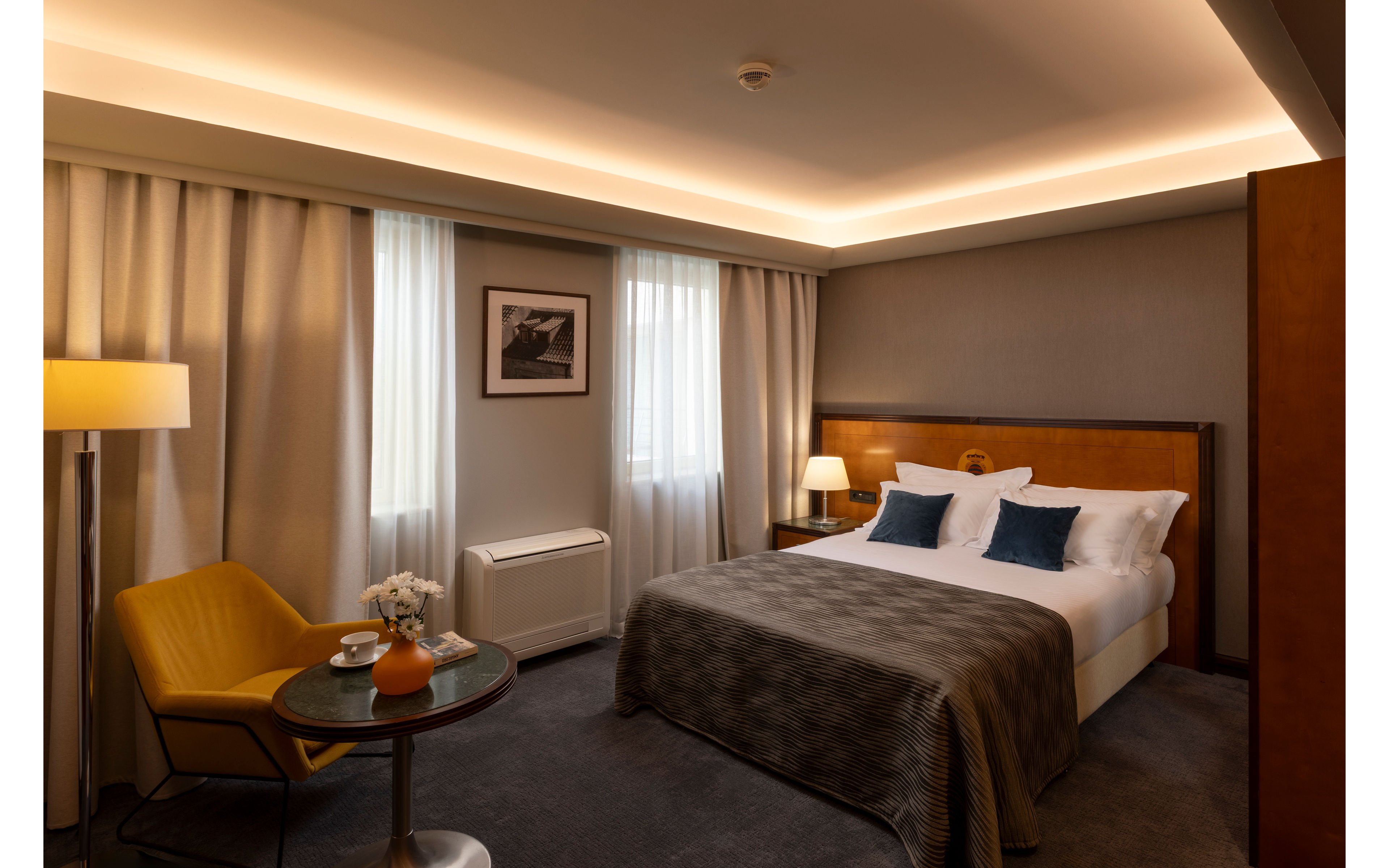 A neatly made double bed with white linens and blue accent pillows sits against a wooden headboard in a modern hotel room. Warm ambient lighting from ceiling fixtures and table lamps creates a welcoming atmosphere. A small round table with chairs, a floor lamp, and a framed black-and-white photograph add to the contemporary decor. Neutral tones, soft curtains, and carpeted flooring contribute to the calm, comfortable setting.