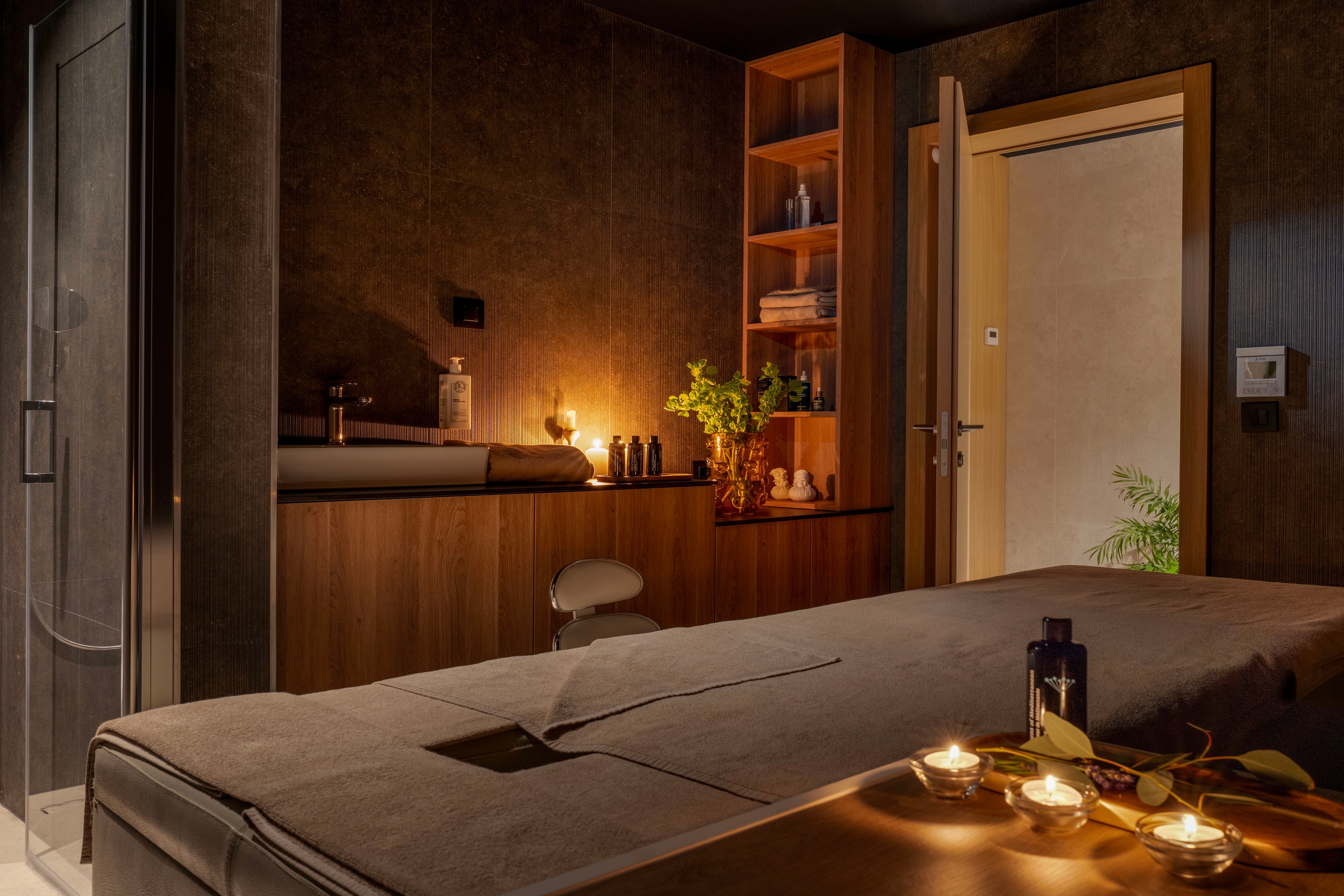 A softly lit spa treatment room features a massage table in the foreground, covered with neutral-toned linens. Warm wooden cabinetry and shelves hold bottles and decor, illuminated by ambient candlelight. A partially open door reveals a plant in the adjacent space, adding a touch of nature to the serene interior. The overall atmosphere is calm and luxurious, with a focus on relaxation and wellness.