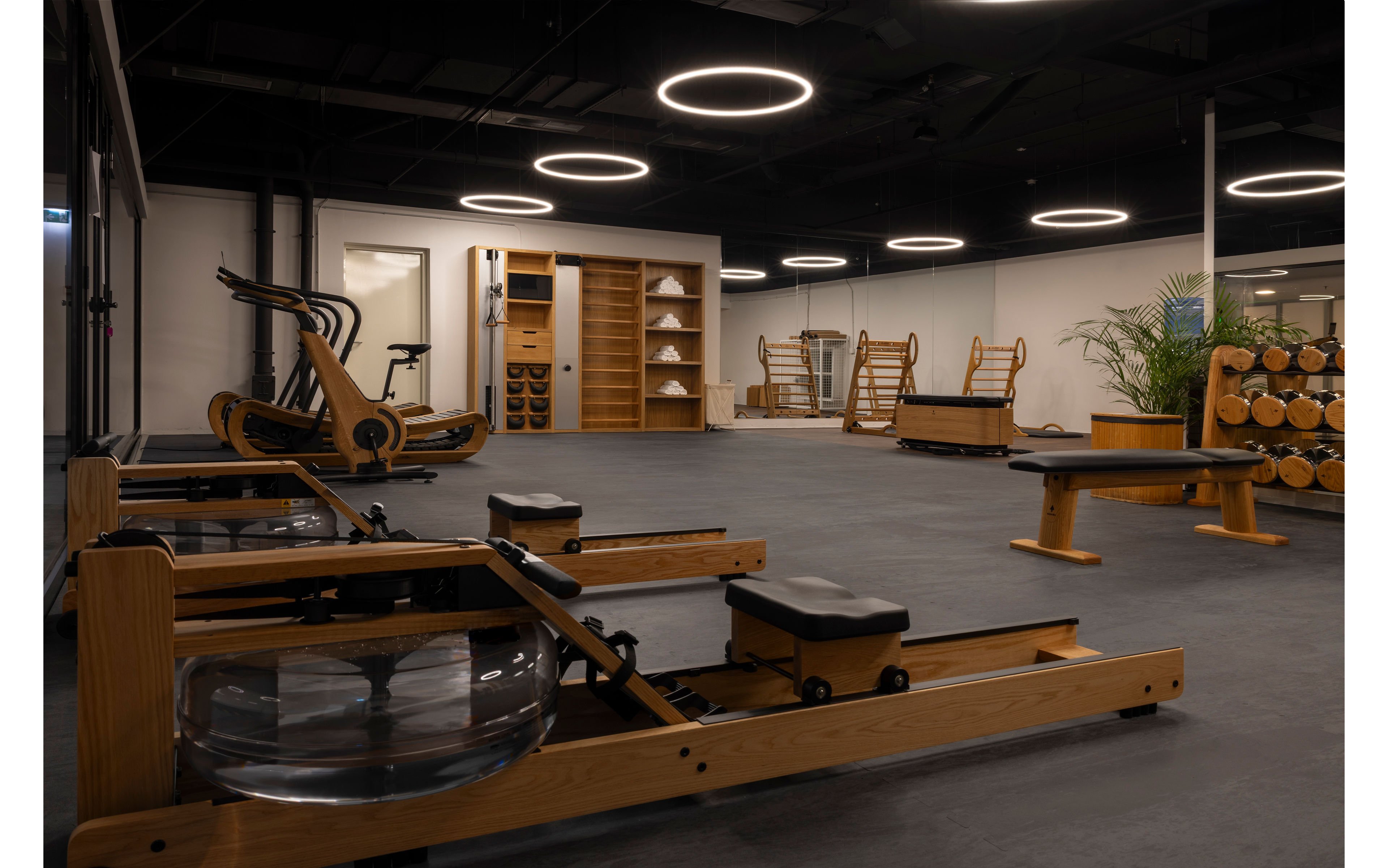 A contemporary fitness studio features several wooden rowing machines and workout benches arranged on a dark floor. Circular ceiling lights create a sleek, modern atmosphere in the spacious room. In the background, wooden storage shelves hold neatly stacked towels and accessories, with additional exercise stations visible. A large plant and wood-accented decor add a warm, natural touch to the minimalist gym interior.