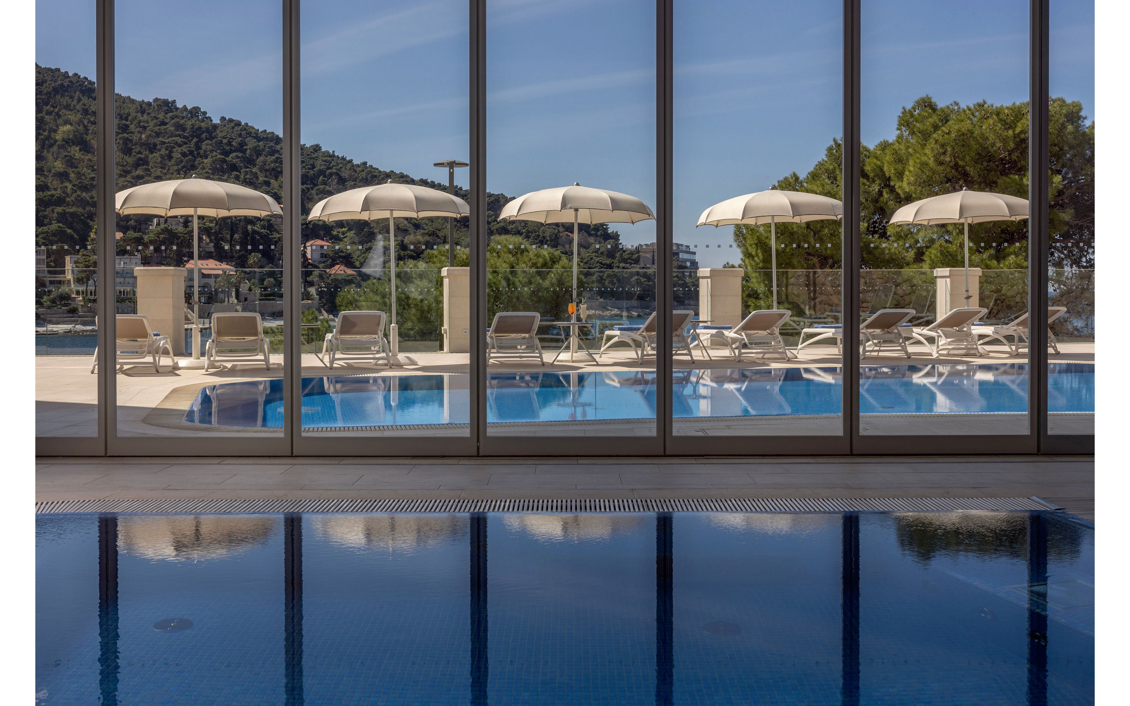A tranquil outdoor pool area is seen through large floor-to-ceiling glass windows. Sun loungers and white umbrellas line the pool deck under clear blue skies. Hills covered with greenery and trees form a scenic backdrop beyond the water. The calm, reflective surface of the indoor pool in the foreground adds to the relaxed vacation atmosphere.