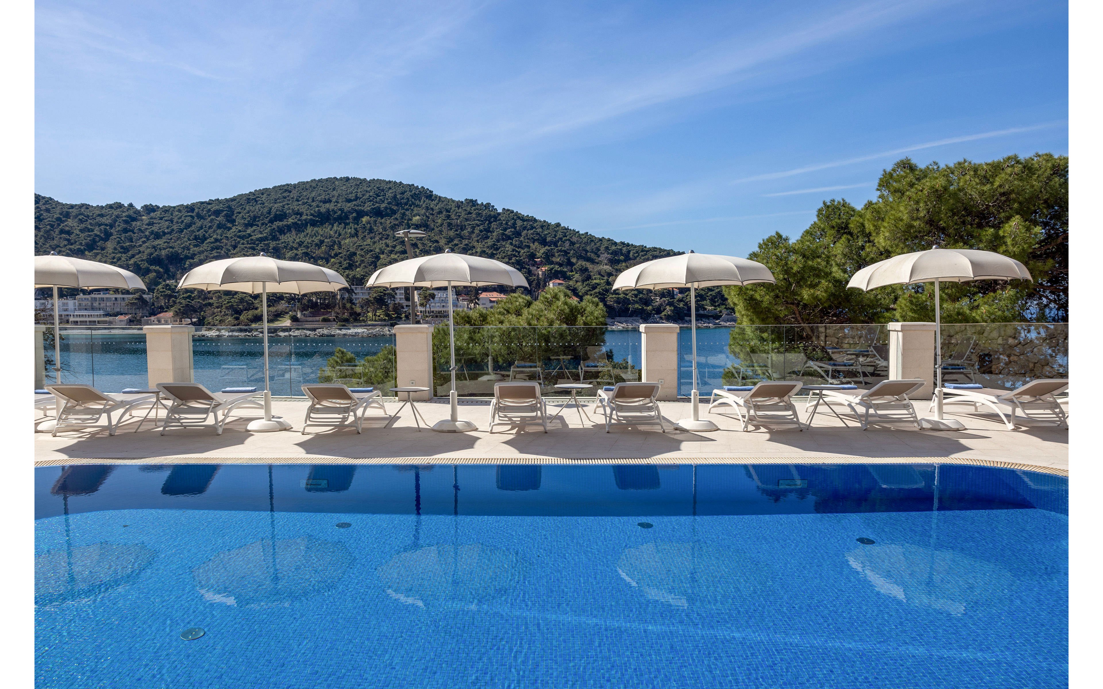A tranquil outdoor swimming pool overlooks a calm blue sea and forested hills. White sun loungers and matching umbrellas line the pool deck in neat rows. Clear glass railings separate the pool area from the water below, enhancing the open coastal view. The bright sky and reflections in the pool create a serene, vacation-like atmosphere.
