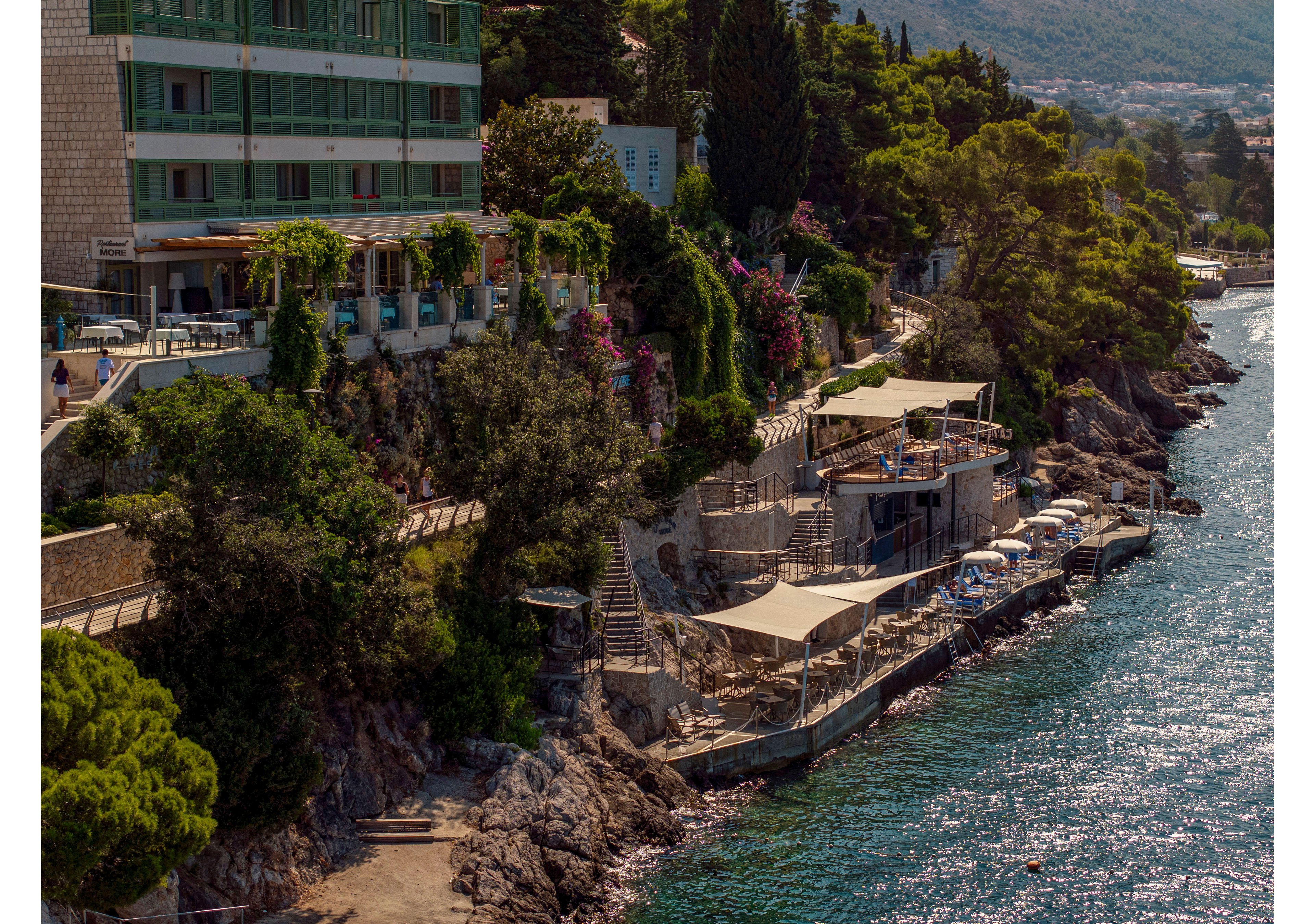 A multi-story seaside hotel stands above a rocky shoreline lined with terraces and sunshades. Stone steps and pathways lead down to waterfront seating areas built into the cliffs. Lush greenery, flowering plants, and tall trees surround the buildings, adding color and texture. Calm blue water borders the scene, with distant hills and town structures visible in the background.