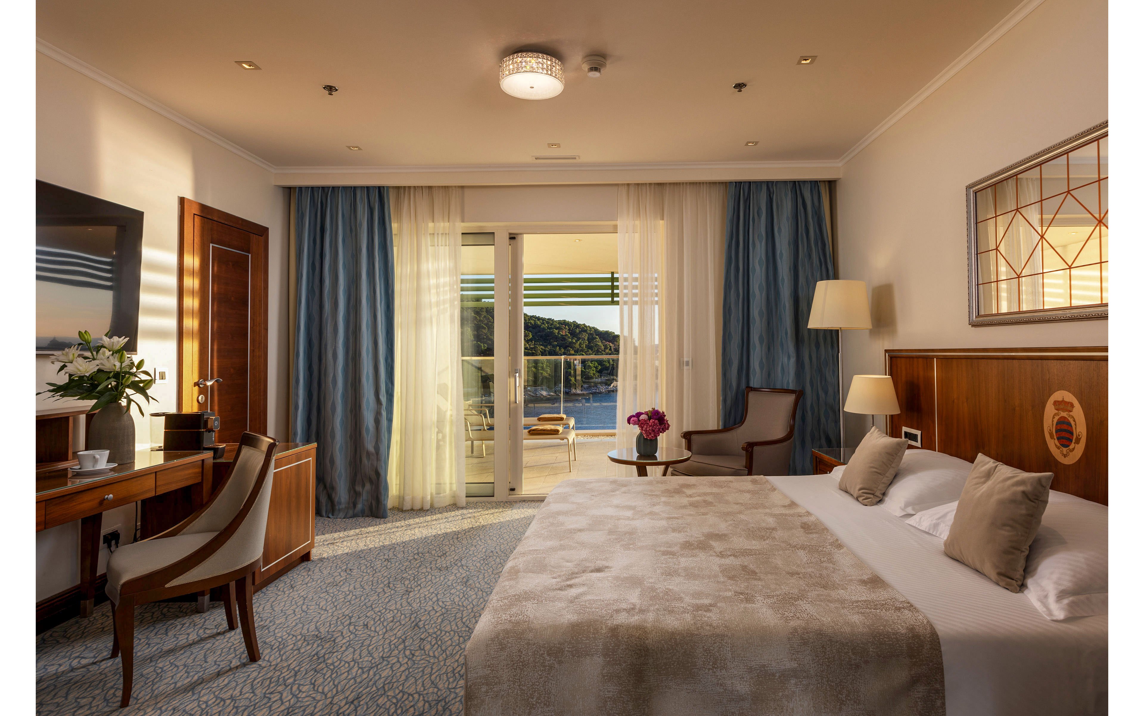 A warmly lit hotel room features a large bed with neutral bedding and decorative pillows. A desk with a chair, television, and flower arrangement sits to the left, while a lounge chair and floor lamp create a cozy corner on the right. Sheer and heavy curtains frame glass doors that open onto a balcony overlooking water and distant hills. Soft lighting, wood furniture, and muted tones give the space a calm, upscale atmosphere.