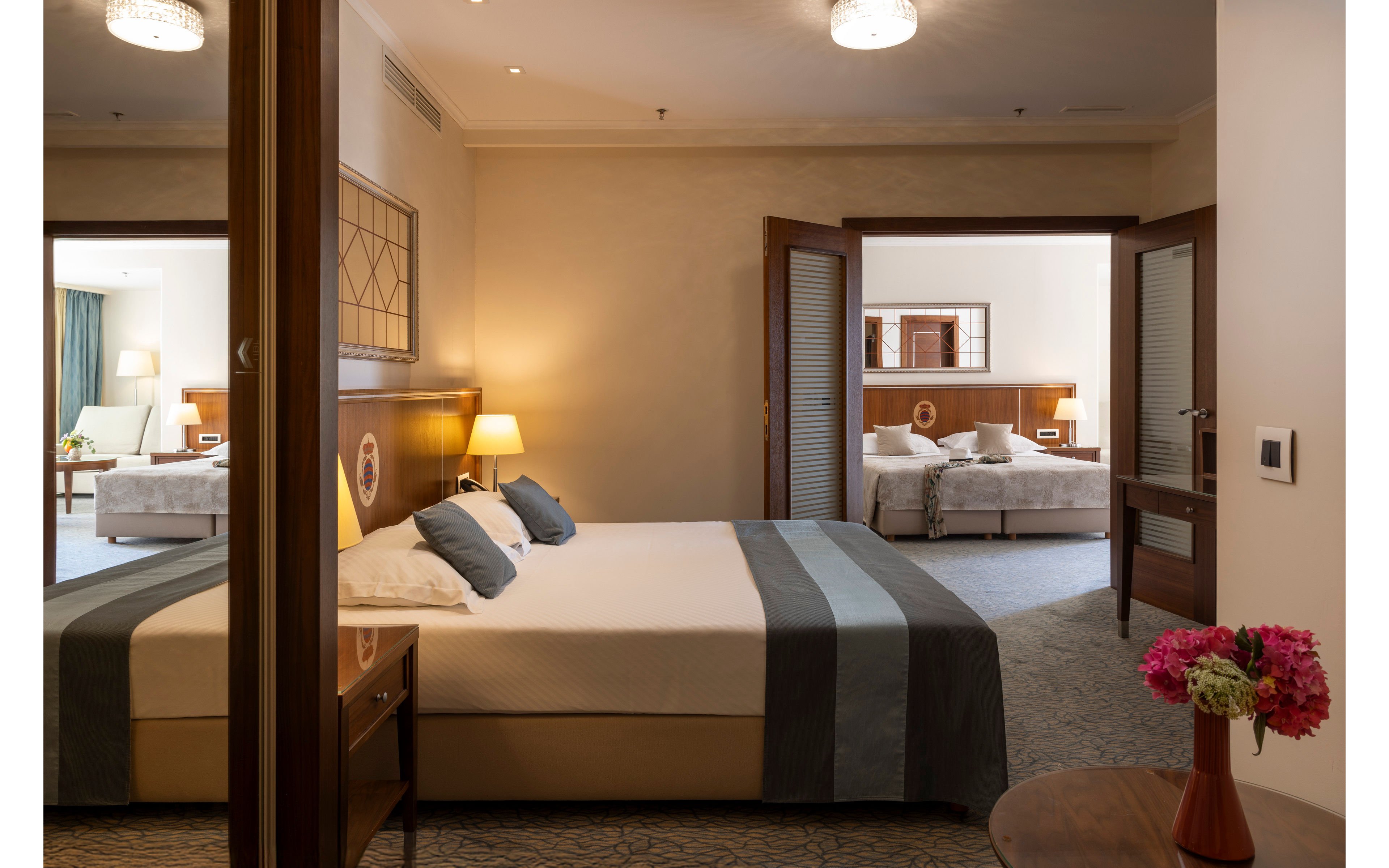 A modern hotel suite features a neatly made double bed with white linens and a gray runner in the foreground. Warm bedside lamps and wooden furniture create a cozy, inviting atmosphere. Two open wooden doors reveal an adjoining room with another bed and soft lighting, suggesting a connected family or group accommodation. A small table with a vase of pink flowers adds a decorative touch to the contemporary interior.