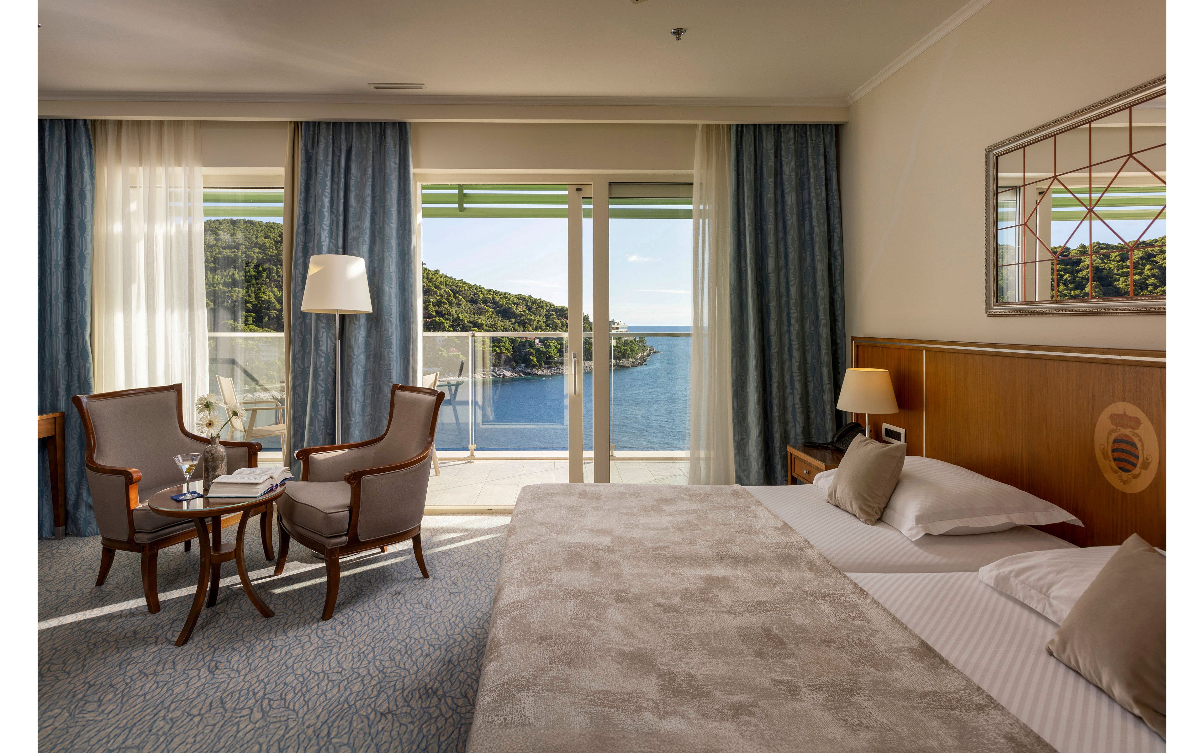 A spacious hotel room features a large bed with neutral bedding and wooden headboard. Two upholstered chairs and a small round table with glassware sit near floor-to-ceiling windows. Sheer and blue curtains frame sliding doors that open to a balcony overlooking a calm sea and green hills. Warm lighting from table lamps and soft colors create a relaxing, upscale atmosphere.