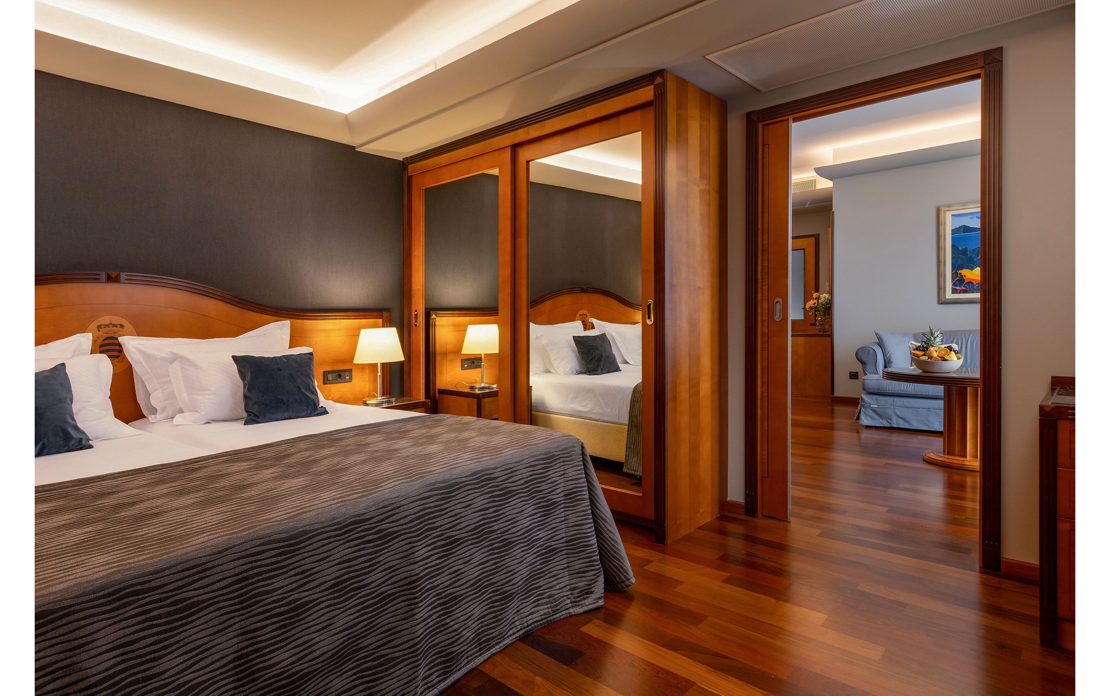 A cozy hotel bedroom features a large bed with striped bedding and dark accent pillows against a wood headboard. Two bedside tables with lamps provide soft, warm lighting that reflects off the polished wooden floor. A mirrored wardrobe door expands the sense of space and shows part of the room’s furnishings. Through an open doorway, a separate sitting area with a sofa, table, and framed artwork is visible, enhancing the suite-like feel.