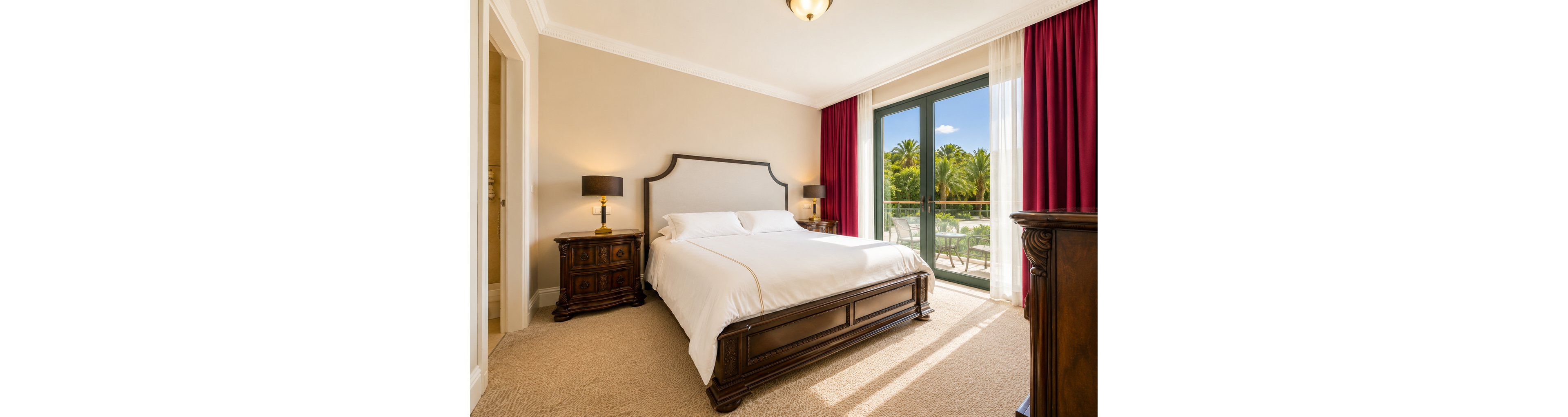 A spacious hotel bedroom features a large wooden bed with crisp white linens centered between two ornate bedside tables. Warm table lamps and neutral walls create a cozy, inviting atmosphere. Floor-to-ceiling glass doors open to a balcony overlooking lush palm trees and a sunny outdoor area. Red curtains frame the doors, adding a touch of color to the otherwise soft, light-toned room.