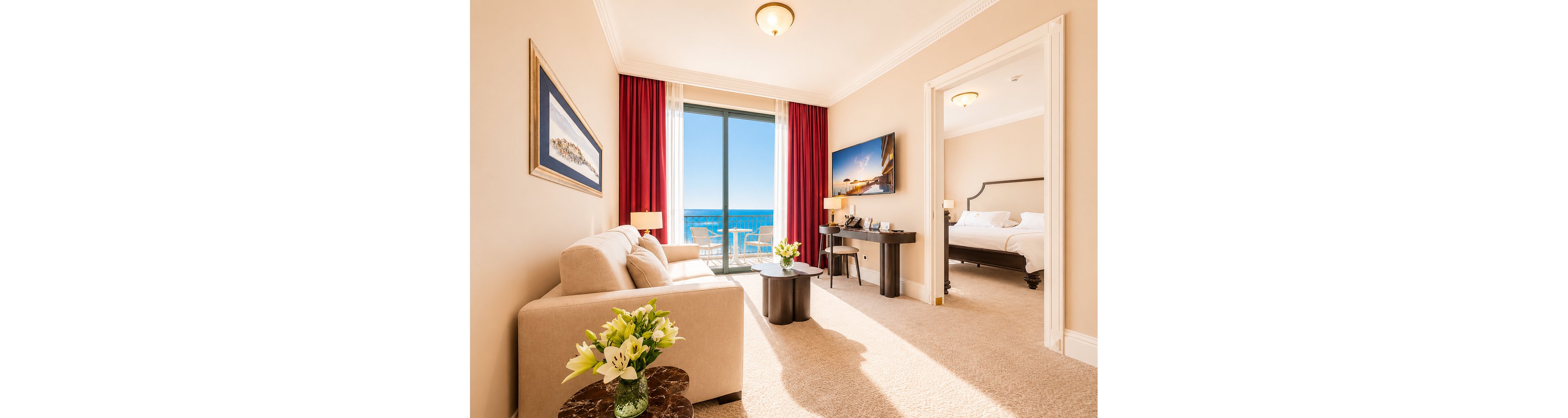 Spacious hotel suite living area with a beige sofa and coffee table is shown in warm natural light. Large glass doors open to a balcony overlooking a bright blue ocean, framed by red curtains. A wall-mounted television, desk, and decorative artwork add modern comfort to the room. Through an open doorway, a neatly made bed is visible in the adjoining bedroom, enhancing the sense of luxury and relaxation.