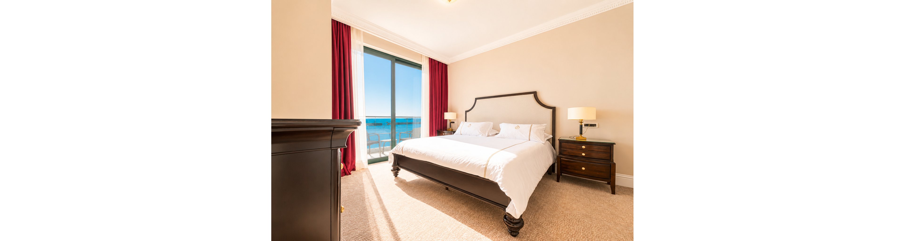A spacious, sunlit bedroom features a large bed with white linens and dark wooden furniture. Floor-to-ceiling glass doors open to a balcony overlooking a clear blue ocean and sky. Red curtains frame the window, contrasting with the beige walls and carpeted floor. The room feels warm and inviting, suggesting a comfortable seaside hotel or resort stay.