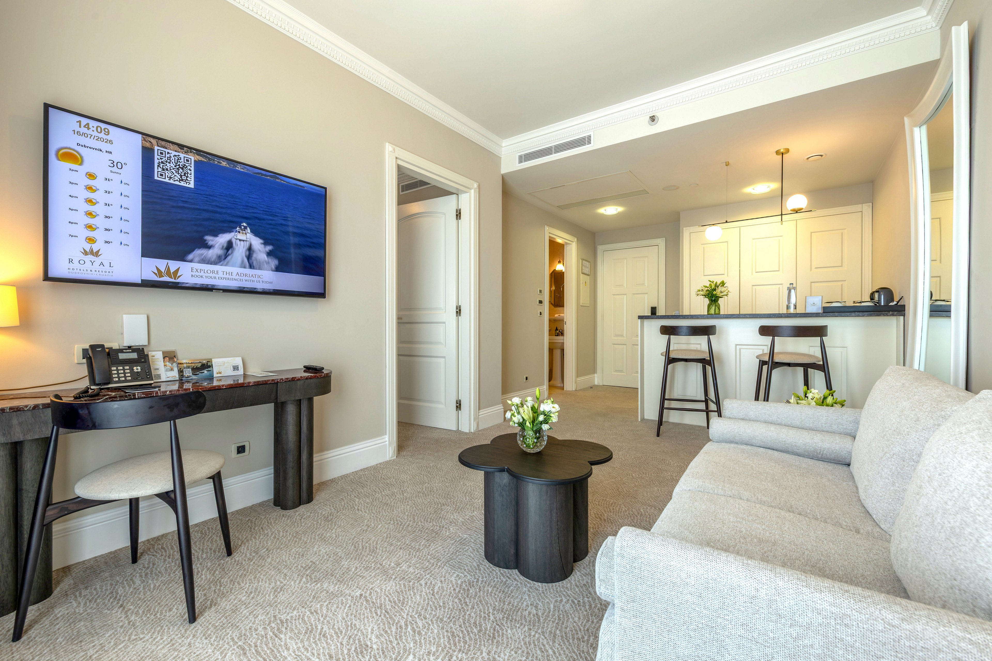 A contemporary hotel suite living area features a sofa facing a wall-mounted flat-screen television. The TV displays a weather forecast panel with temperatures and a QR code beside an ocean scene. A compact kitchenette with bar stools and white cabinetry is visible in the background. Neutral beige walls and carpet, along with dark wood furniture and fresh flowers, create a clean and inviting atmosphere.
