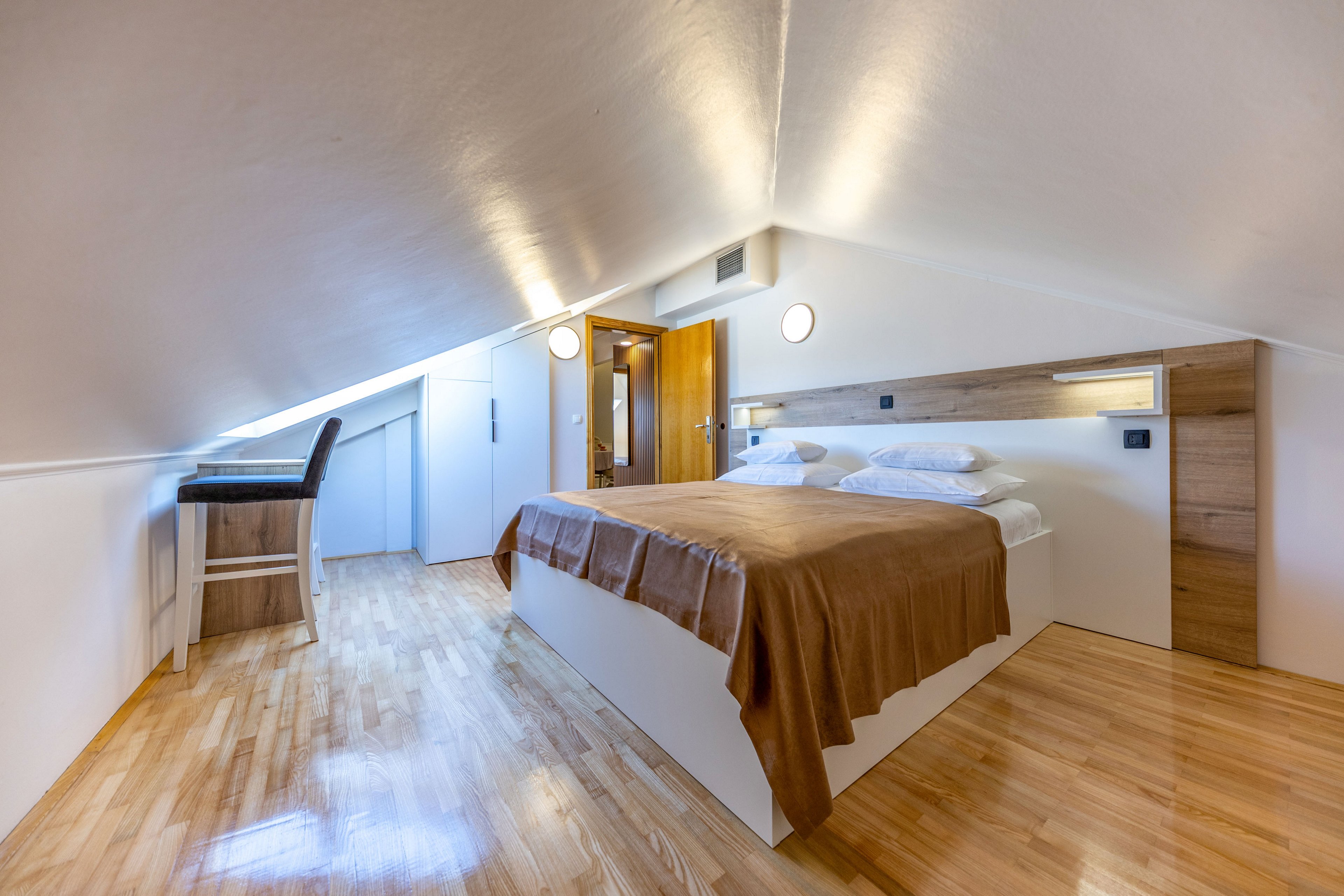 Bright attic bedroom featuring a double bed with brown bedding and white pillows. The space has sloped ceilings, light wooden floors, and built-in storage cabinets along the wall. A small bar-height table with two stools sits near the entrance, adding a functional touch. Warm lighting and minimalist decor create a cozy, contemporary atmosphere.