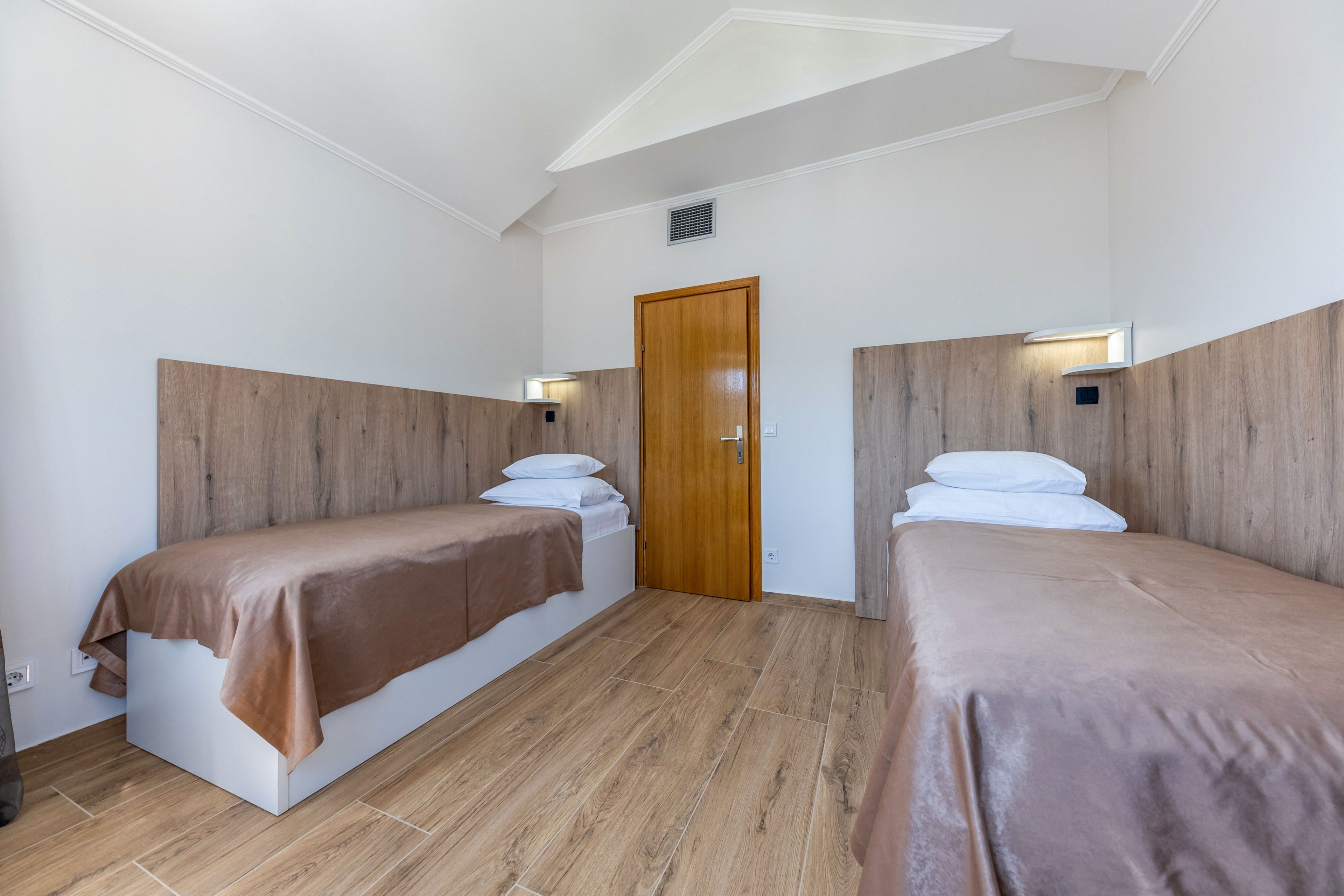 A bright, minimalist bedroom features two single beds with brown bedspreads and white pillows. The room has light wood paneling behind the beds and matching wooden flooring. A simple wooden door is centered on the back wall, with small wall-mounted lights above each bed. The overall design is clean, modern, and uncluttered, emphasizing neutral tones and natural materials.