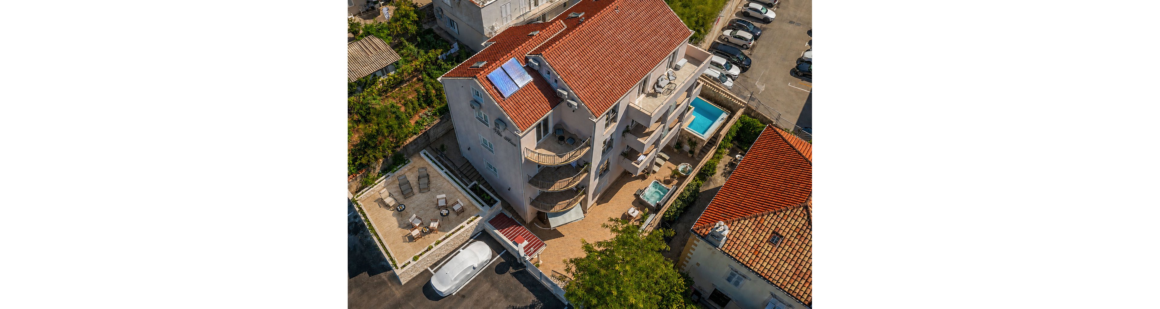 An aerial view shows a multi-story apartment building with a red-tiled roof in a Mediterranean setting. The property features several balconies, a small swimming pool, and an outdoor hot tub on the ground level terrace. Adjacent to the building is a paved patio area with lounge chairs and tables, as well as a parked white vehicle nearby. Surrounding structures and a parking area indicate a residential neighborhood close to a hillside garden.